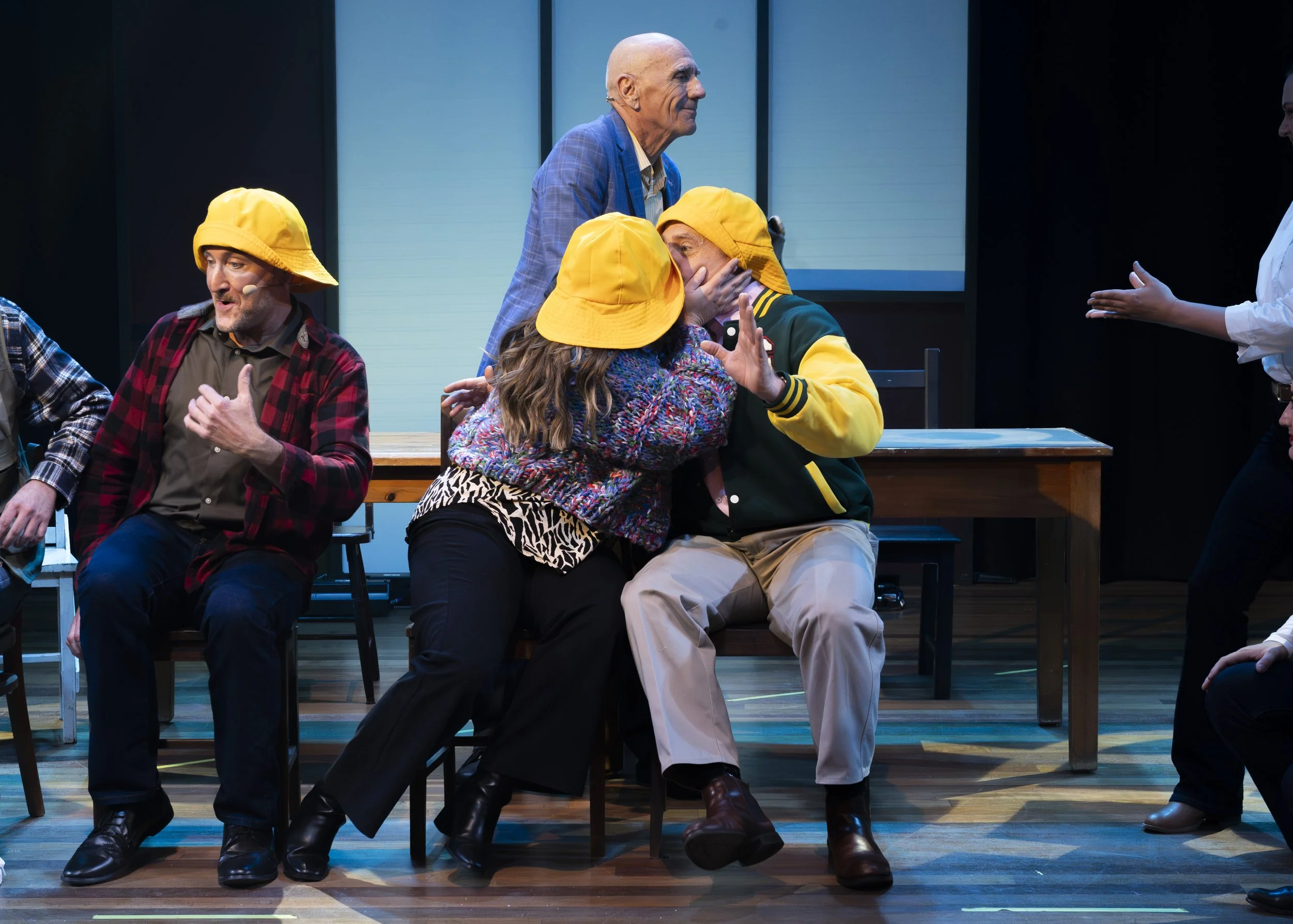 A theatrical stage scene with five actors, some wearing yellow hats, engaged in a comedic or dramatic moment. One actor in a blue jacket is standing behind two characters sitting on a bench, with one of these characters covering their face in surpris