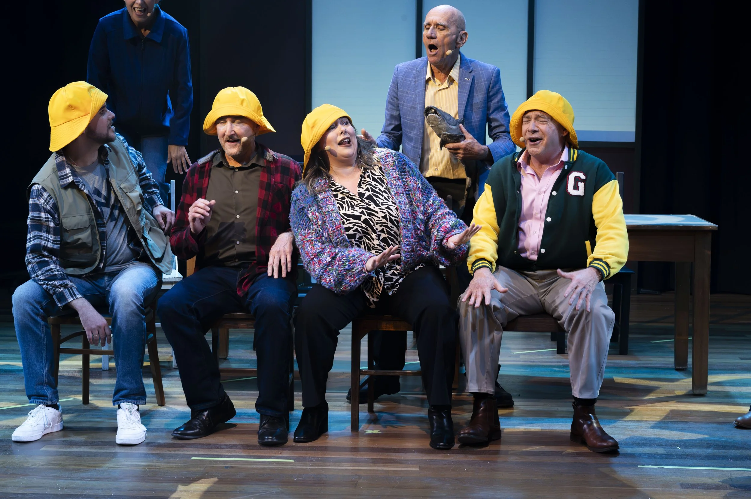 A theatrical stage scene with seven actors, four wearing yellow hats, engaged in a comedic or dramatic performance with expressive faces and gestures.