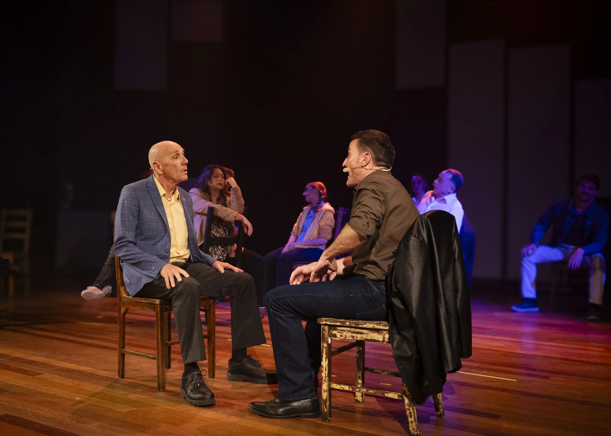 A stage scene from a play shows two men sitting on wooden chairs facing each other, engaging in a conversation. The man on the left is bald, wearing a blue plaid blazer and black pants, with a surprised or concerned expression. The man on the right h