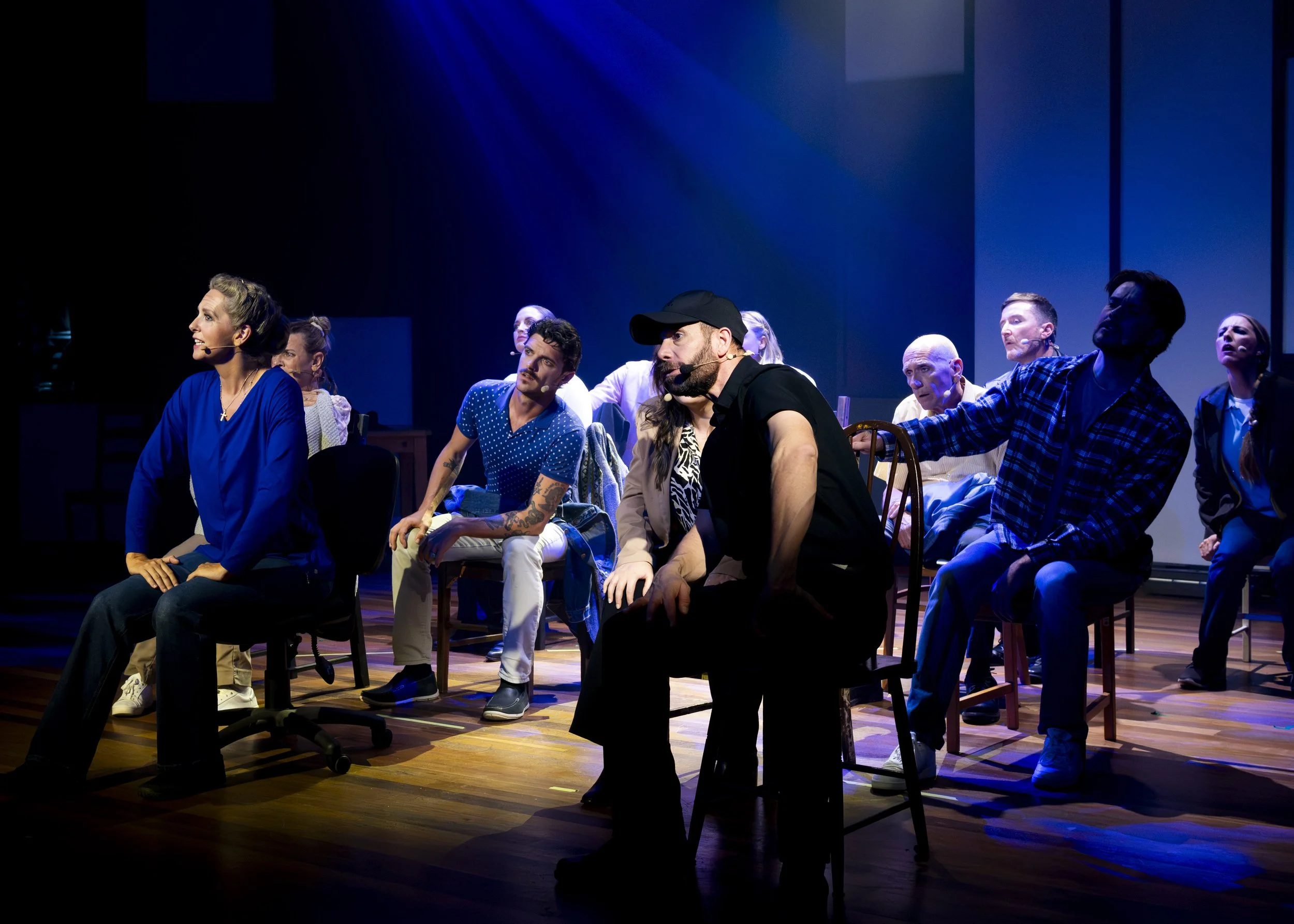 People participating in a stage performance or rehearsal, sitting on chairs on a wooden floor, with dramatic lighting and a dark background.