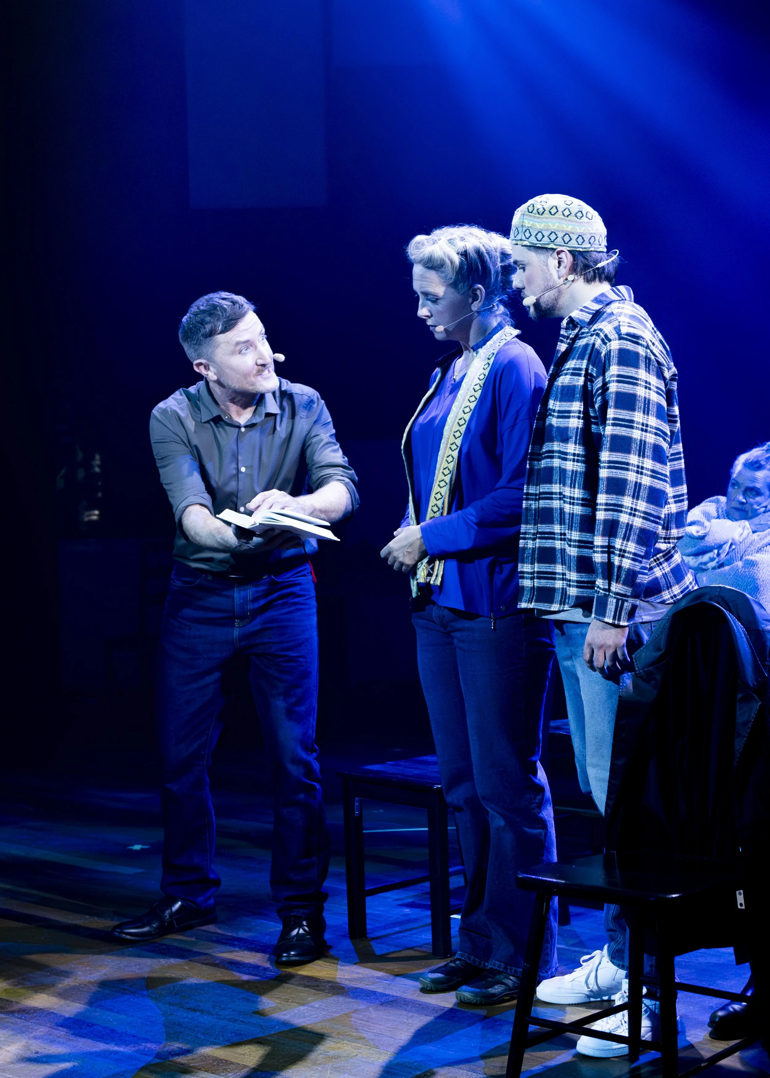 Three actors on stage under blue lighting, engaged in a play. One man is holding a script or book, speaking to a woman and a man. The woman and man are listening attentively. Background includes a seated woman partially visible.