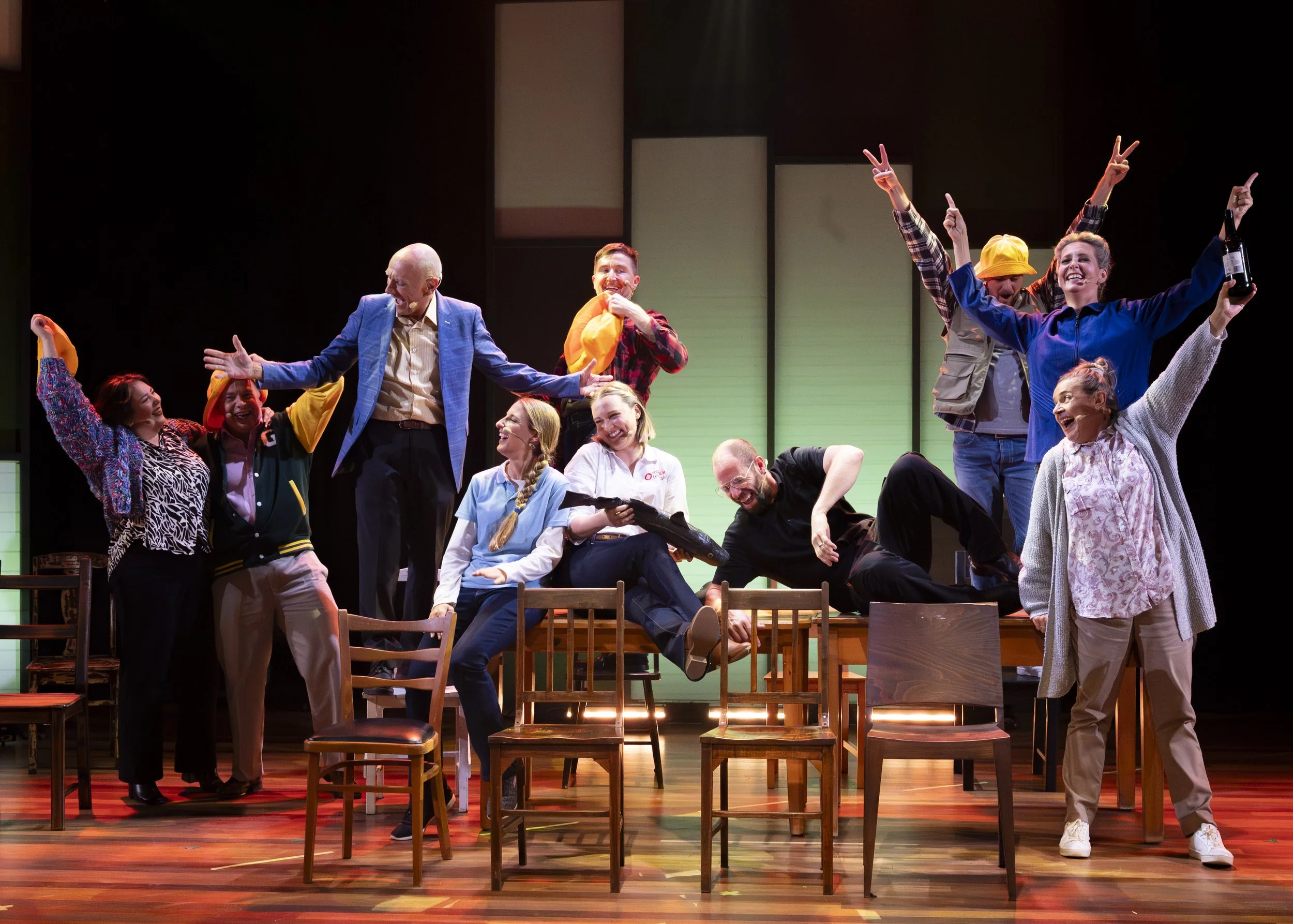 Theater group performing on stage with multiple actors smiling and posing, some sitting on chairs and others standing or leaning on tables, with vibrant lighting