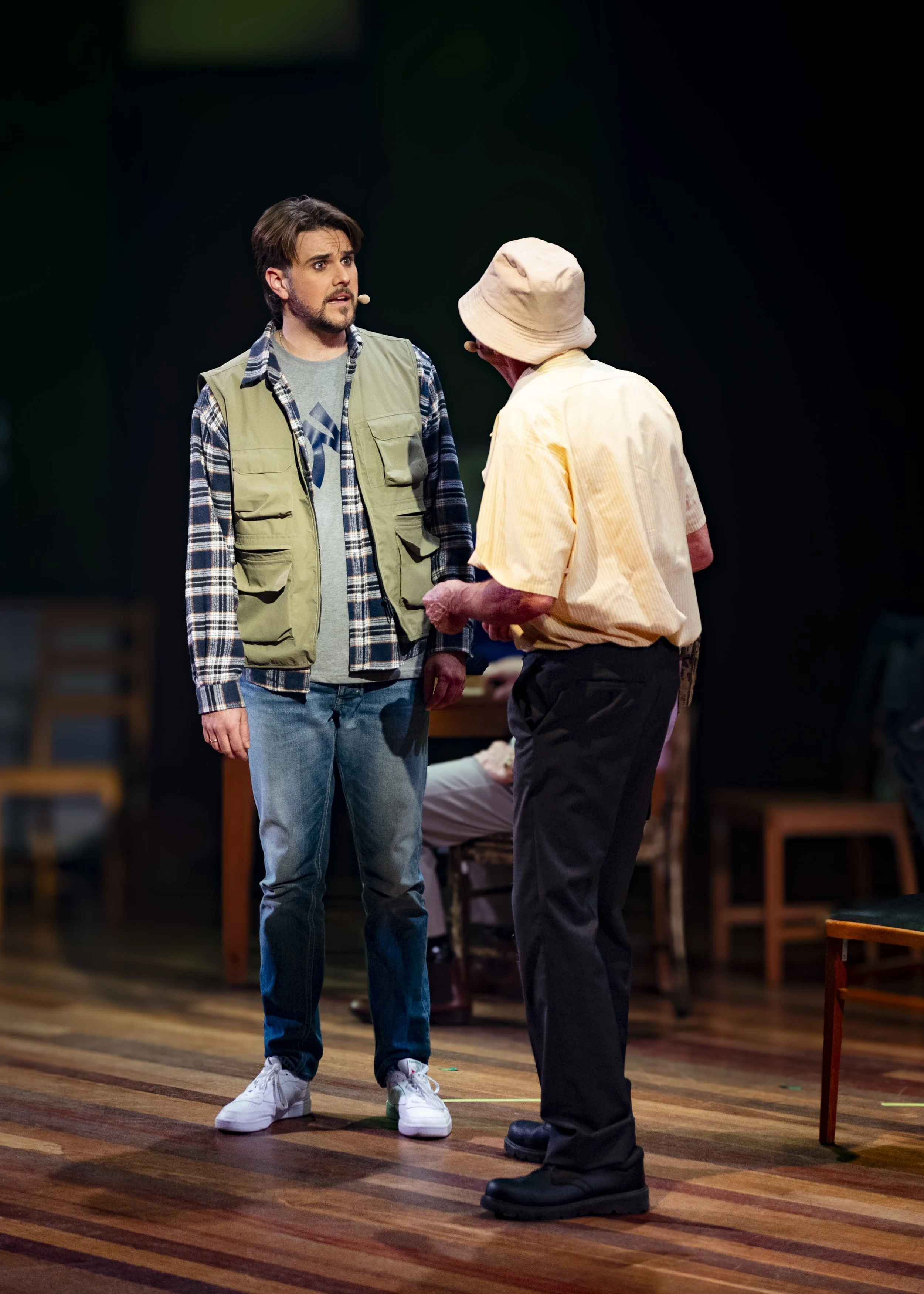 Two men talking on a stage, one wearing a plaid shirt and vest, the other in a yellow shirt and bucket hat.