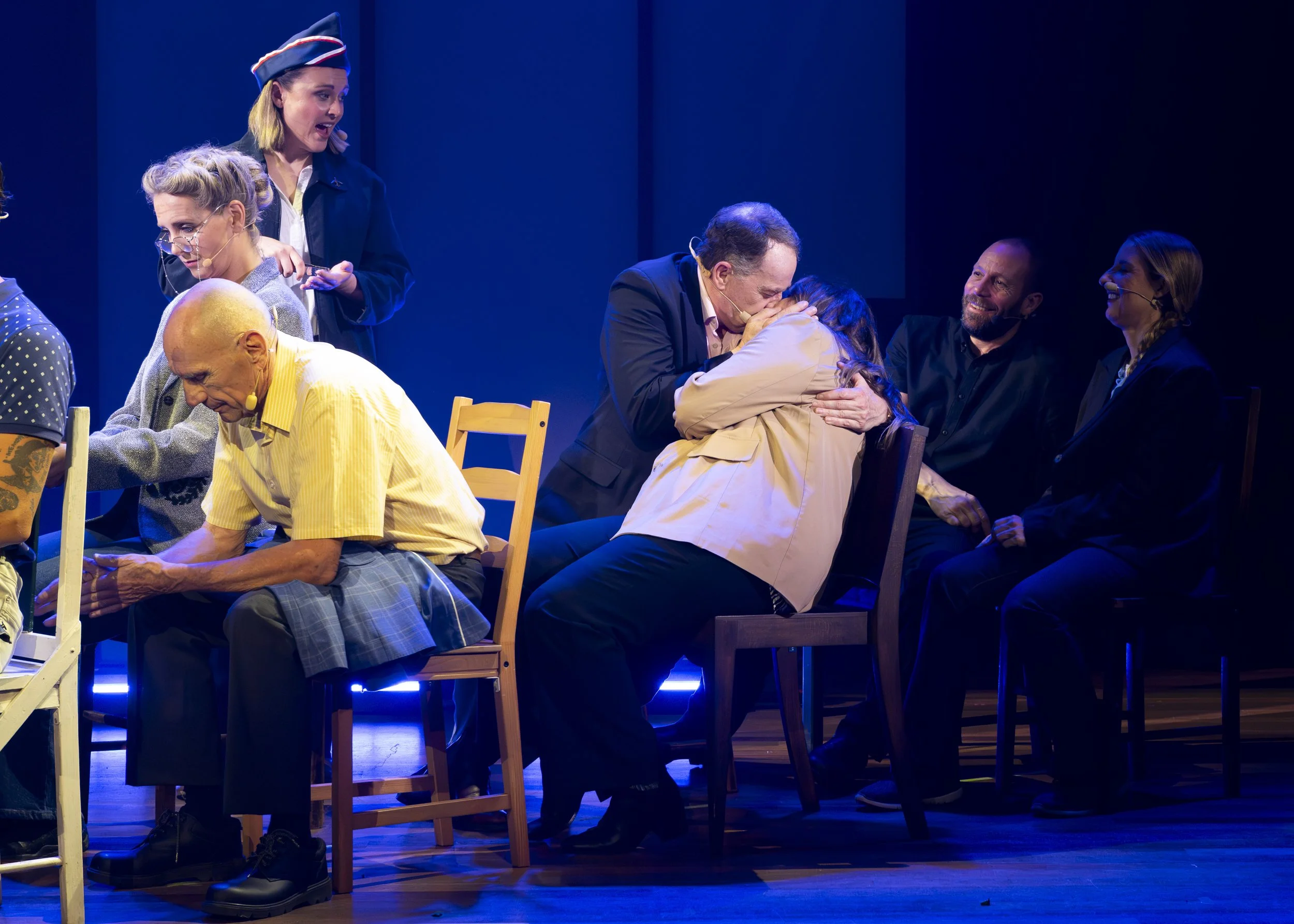 People on stage, some hugging, others sitting and observing, in a theatrical performance with blue lighting.
