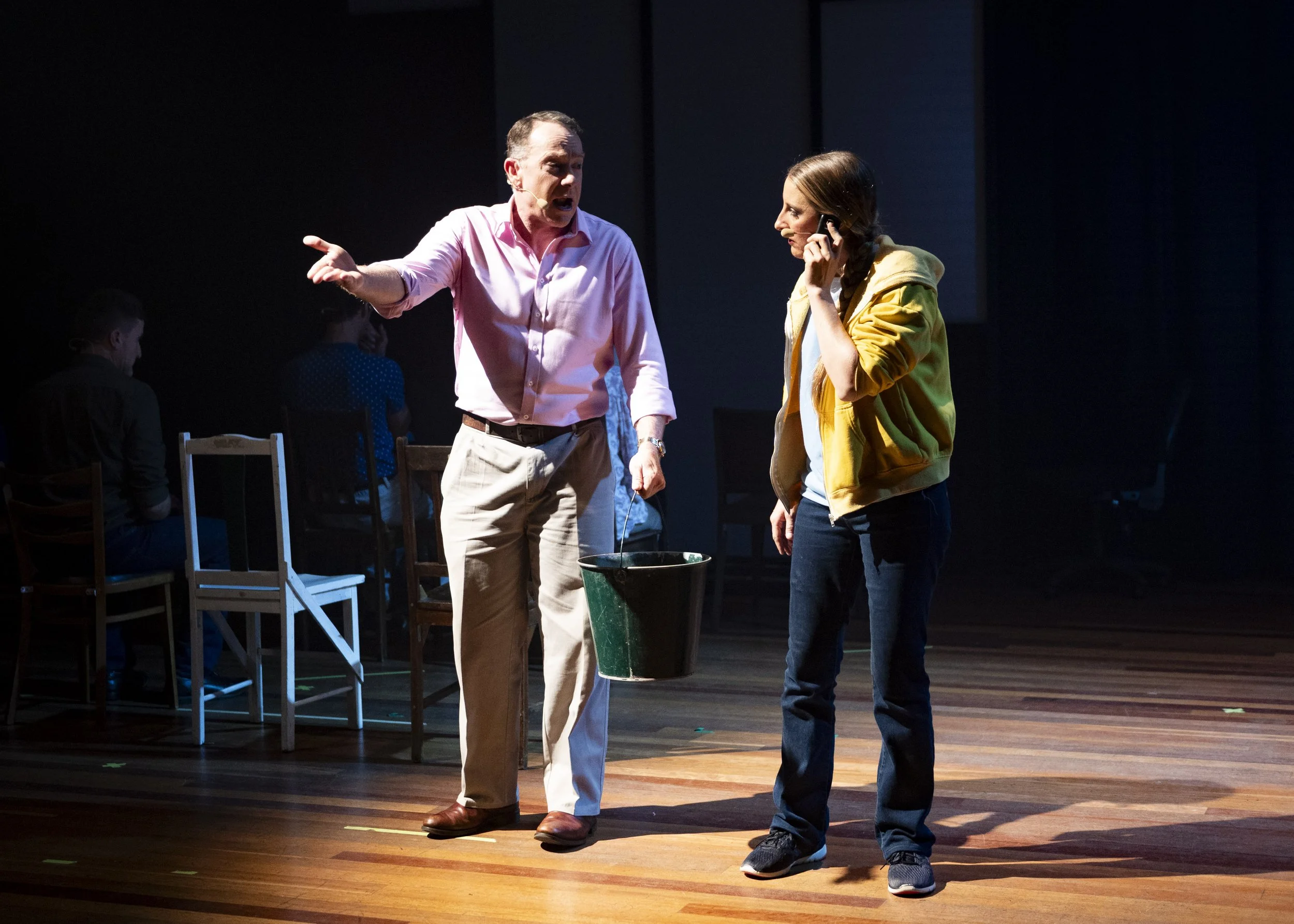 A man in a pink shirt and beige pants gesturing and speaking to a woman in a yellow hoodie and dark jeans, who is on the phone. They are on a stage with a wooden floor, with three chairs and two people seated in the background.