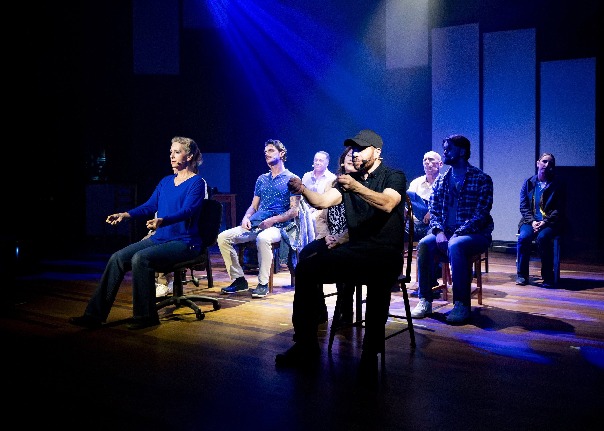 A group of seven people on stage participating in a performance or discussion, with a man in a black shirt and cap speaking into a headset microphone and gesturing.