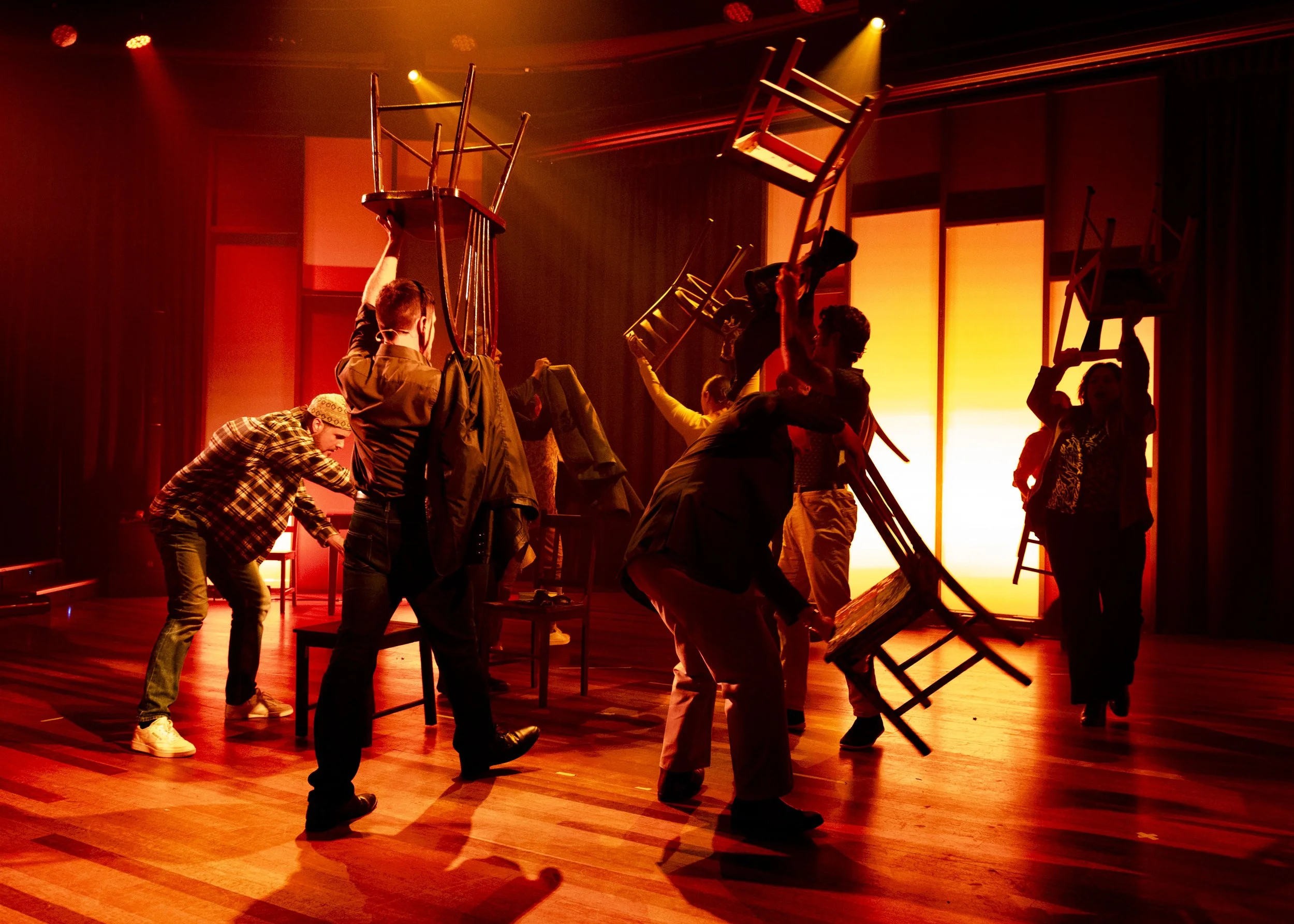 People on stage, lifting chairs, in a warm, orange lighting setting, possibly rehearsing or preparing for a performance.
