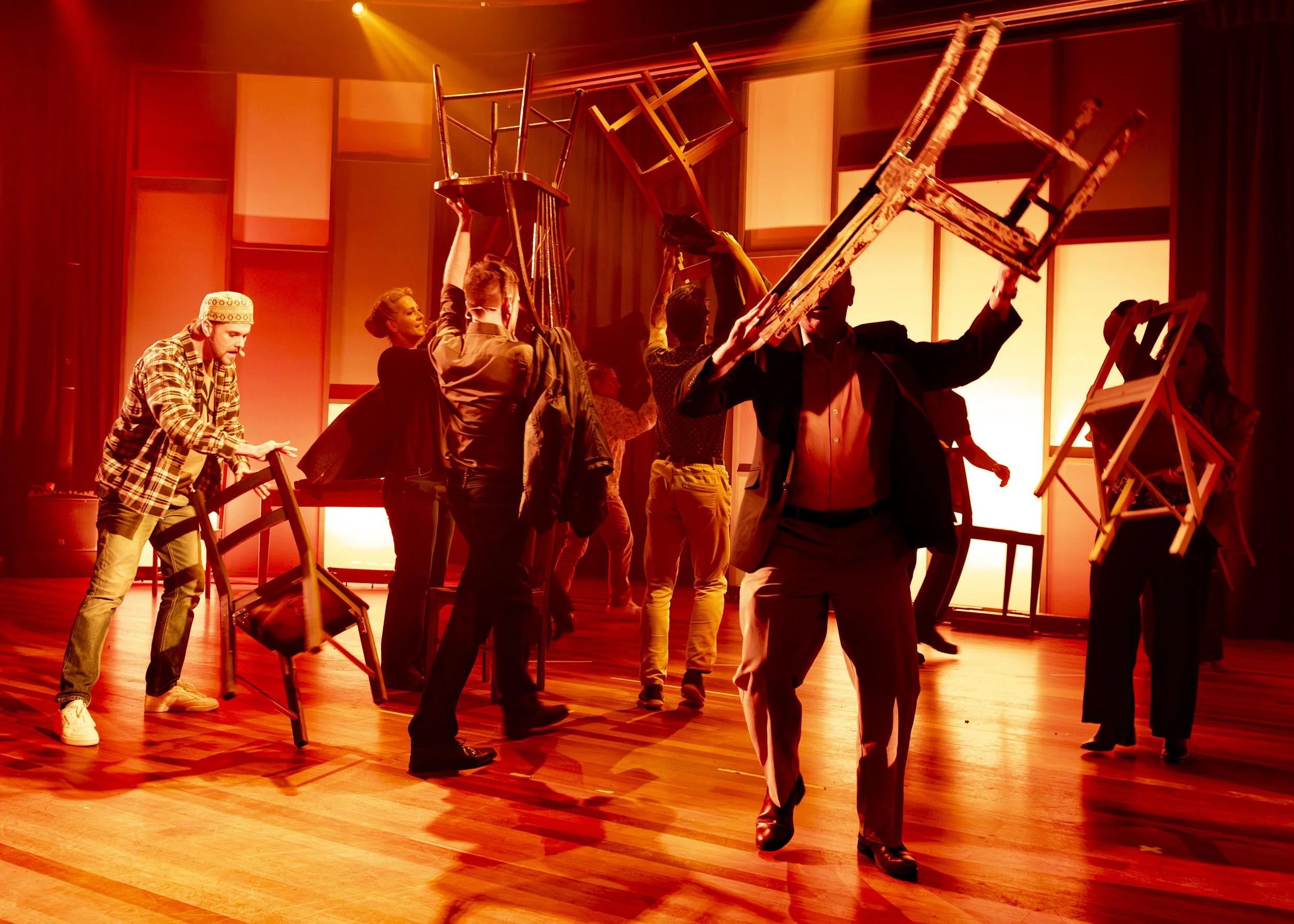 People on stage balancing chairs during a rehearsal or performance, with warm lighting and a wooden floor.