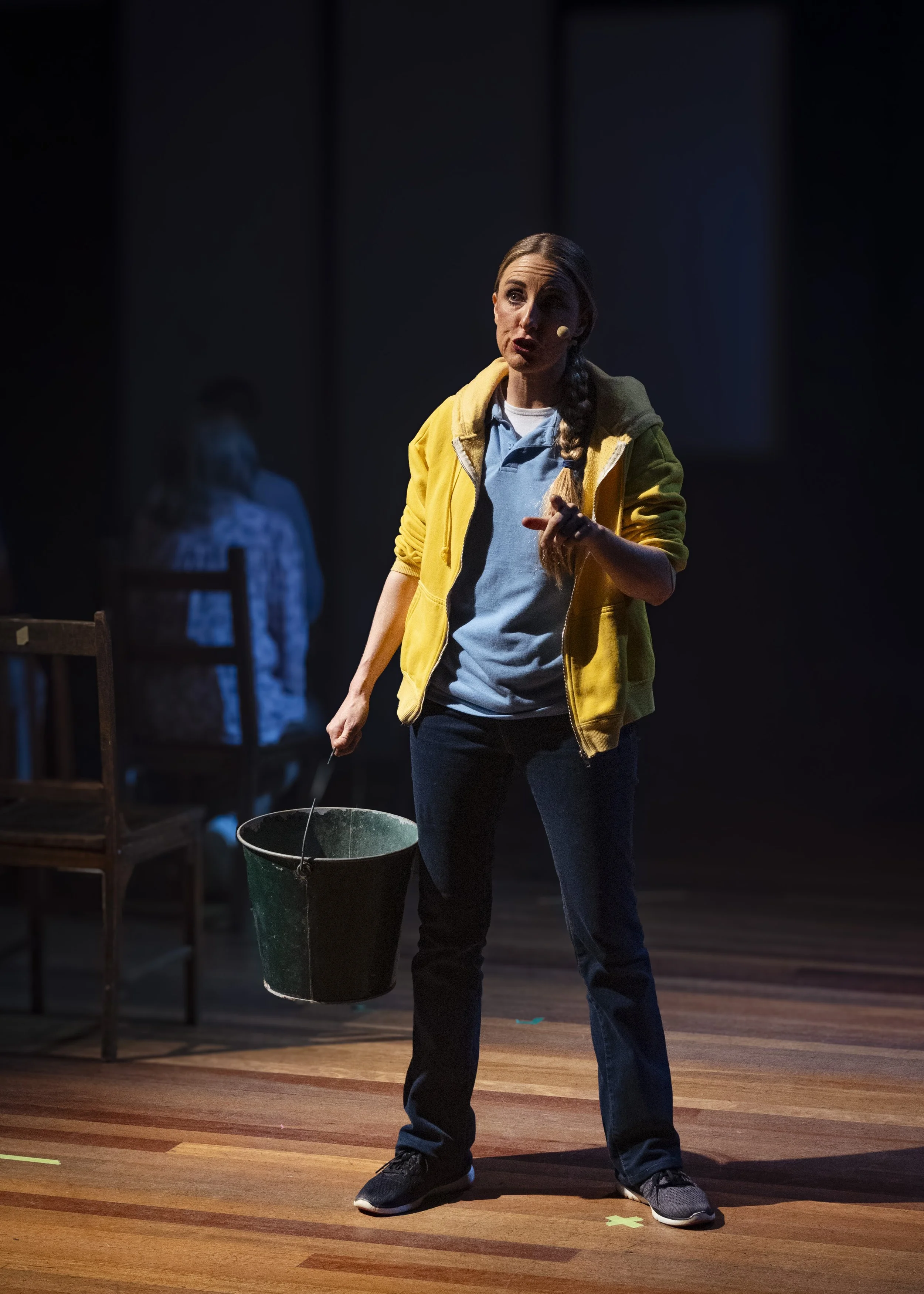 Woman in yellow jacket standing on stage with a microphone headset, holding a bucket, with a person sitting in the background.