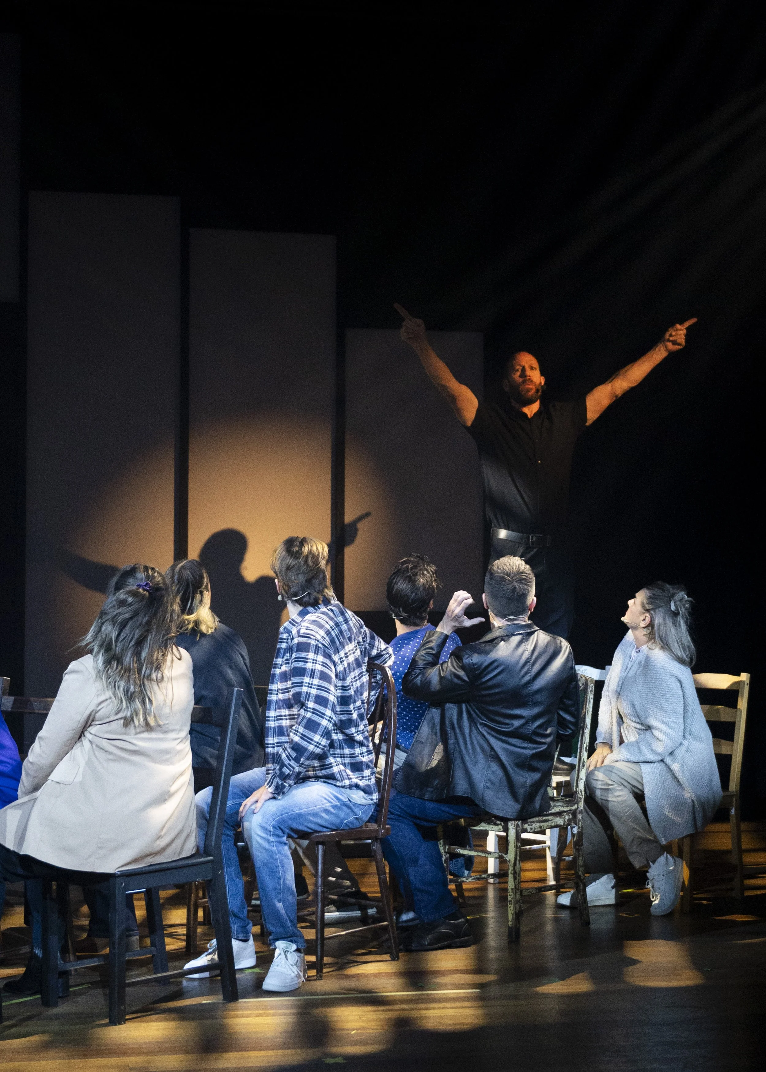 A man stands on stage with arms raised, gesturing outward, as a group of people sit in front of him, listening and engaging during a theatrical performance.