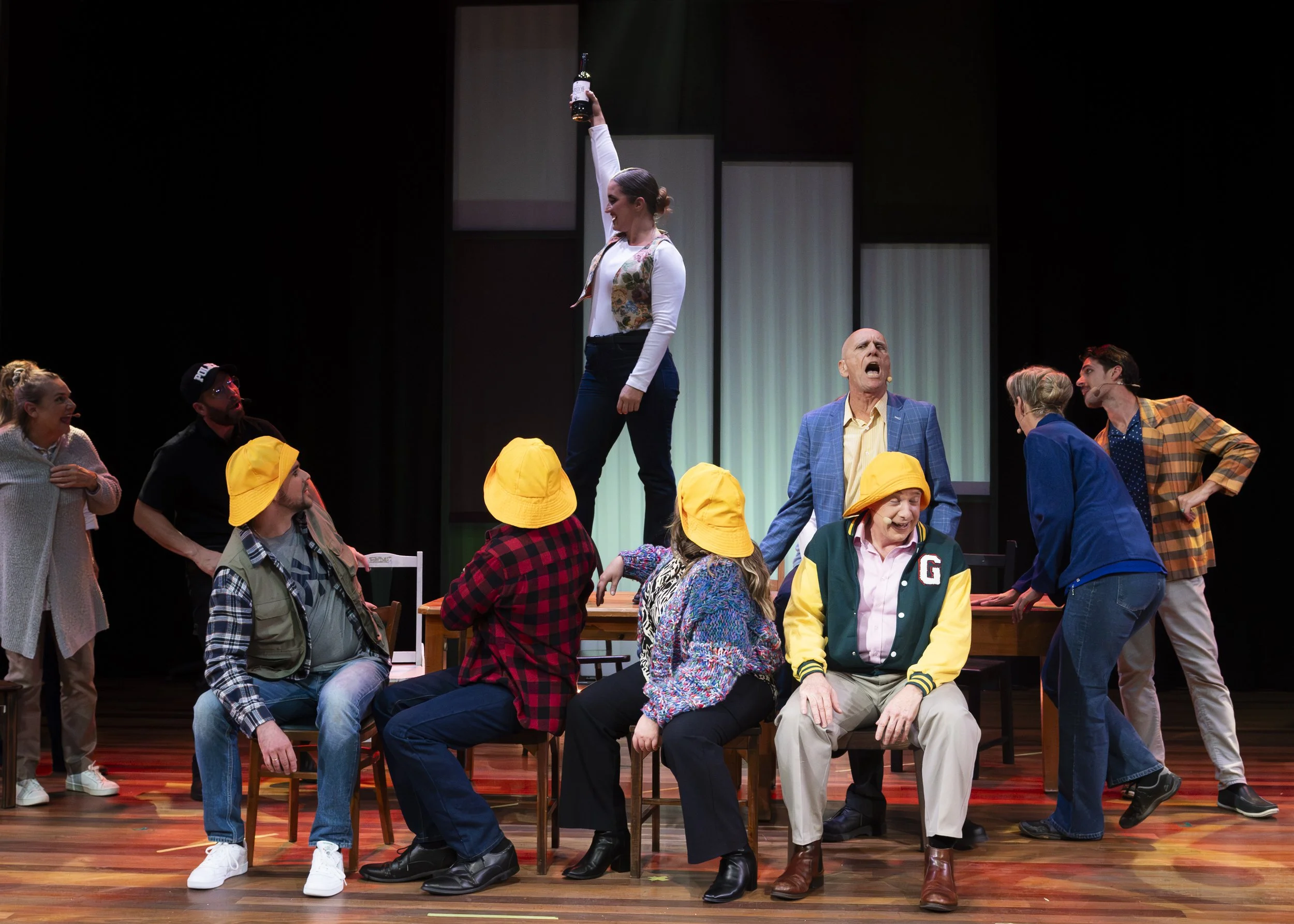 Theatrical performance with multiple actors on stage, one woman standing with a bottle raised, others seated or standing around a table, wearing colorful costumes and some with yellow hats, expressive faces, in a theatrical set with a dark background