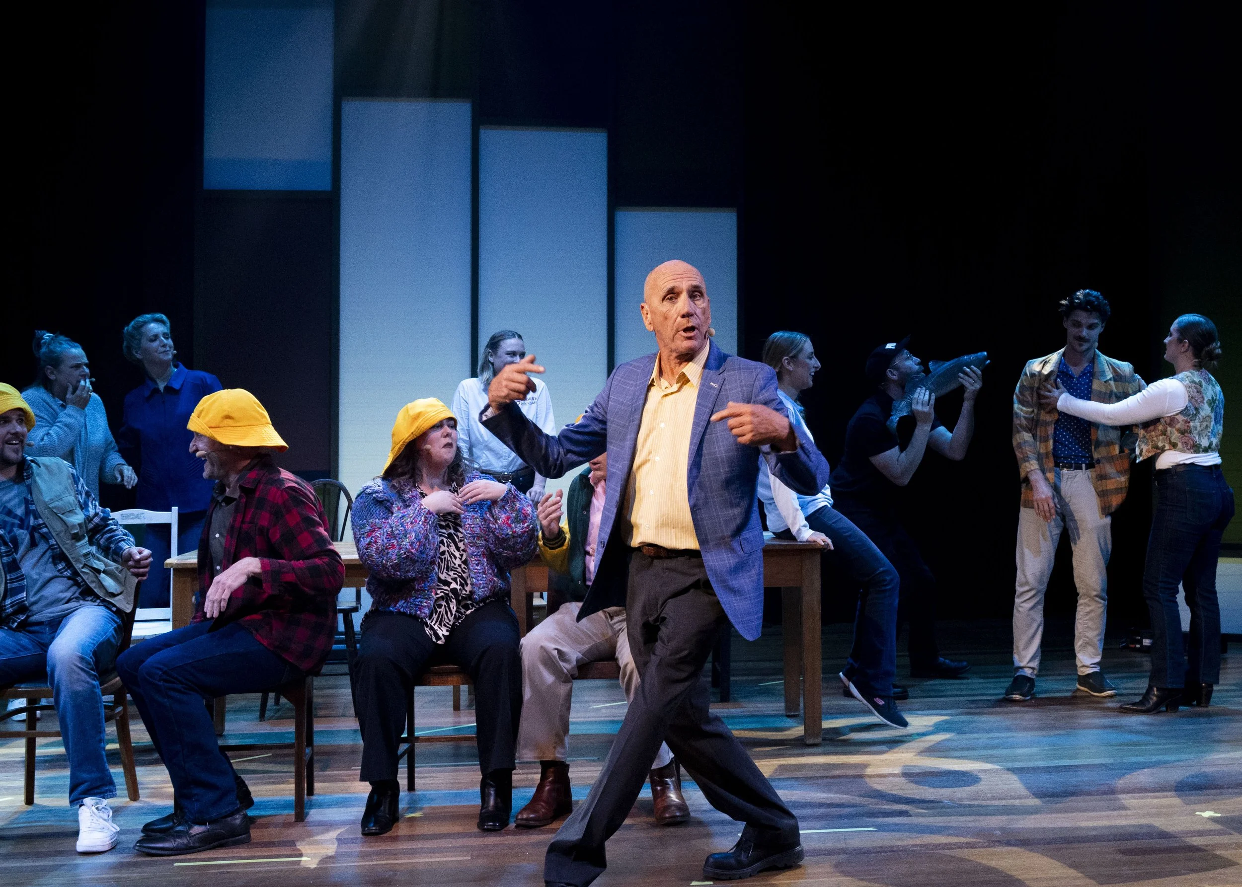The stage of a theatrical play with actors portraying various characters, some seated and some standing, with a man in a checkered blazer and yellow shirt speaking and gesturing at the center.