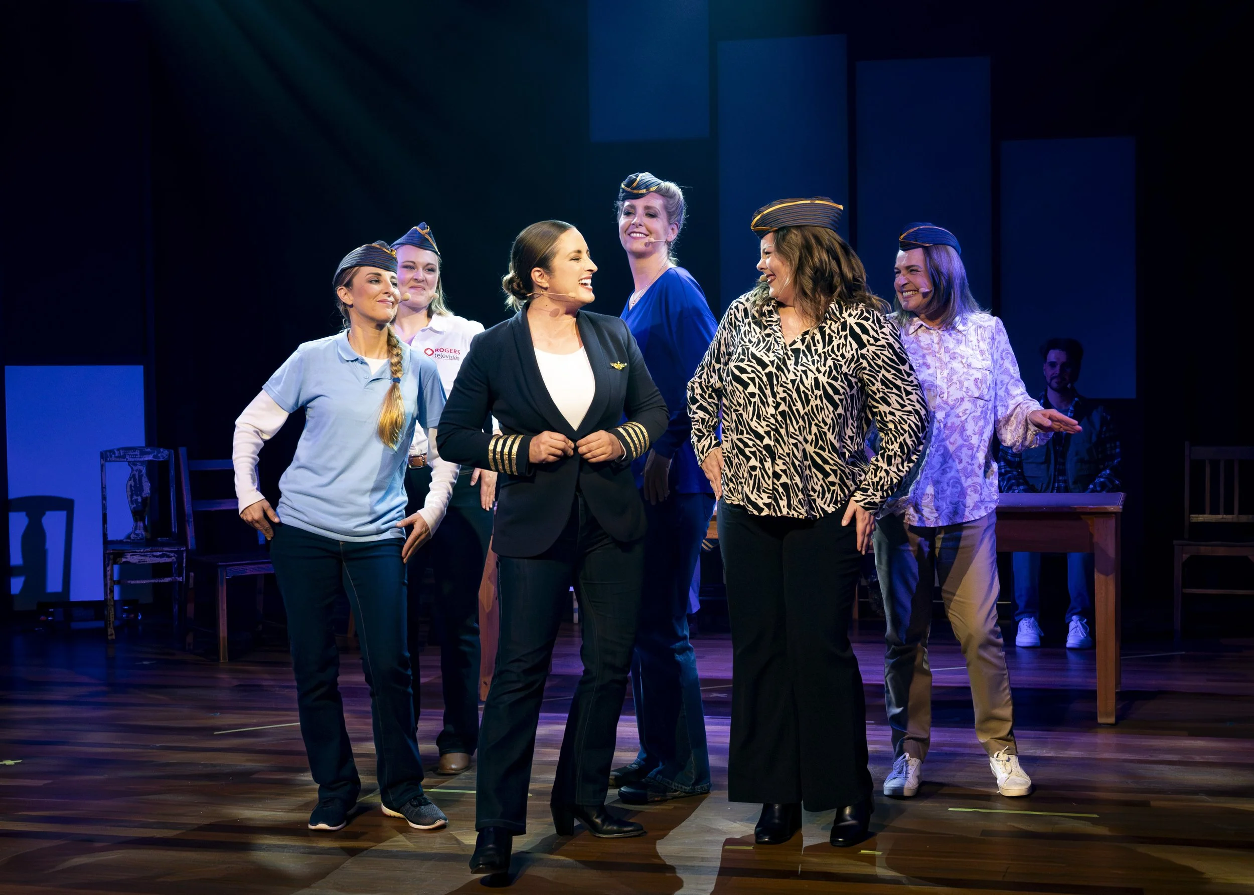 Actors on stage in a theatrical production, some wearing pilot hats, engaged in a scene with smiles and conversation, with a dark background and stage lighting.