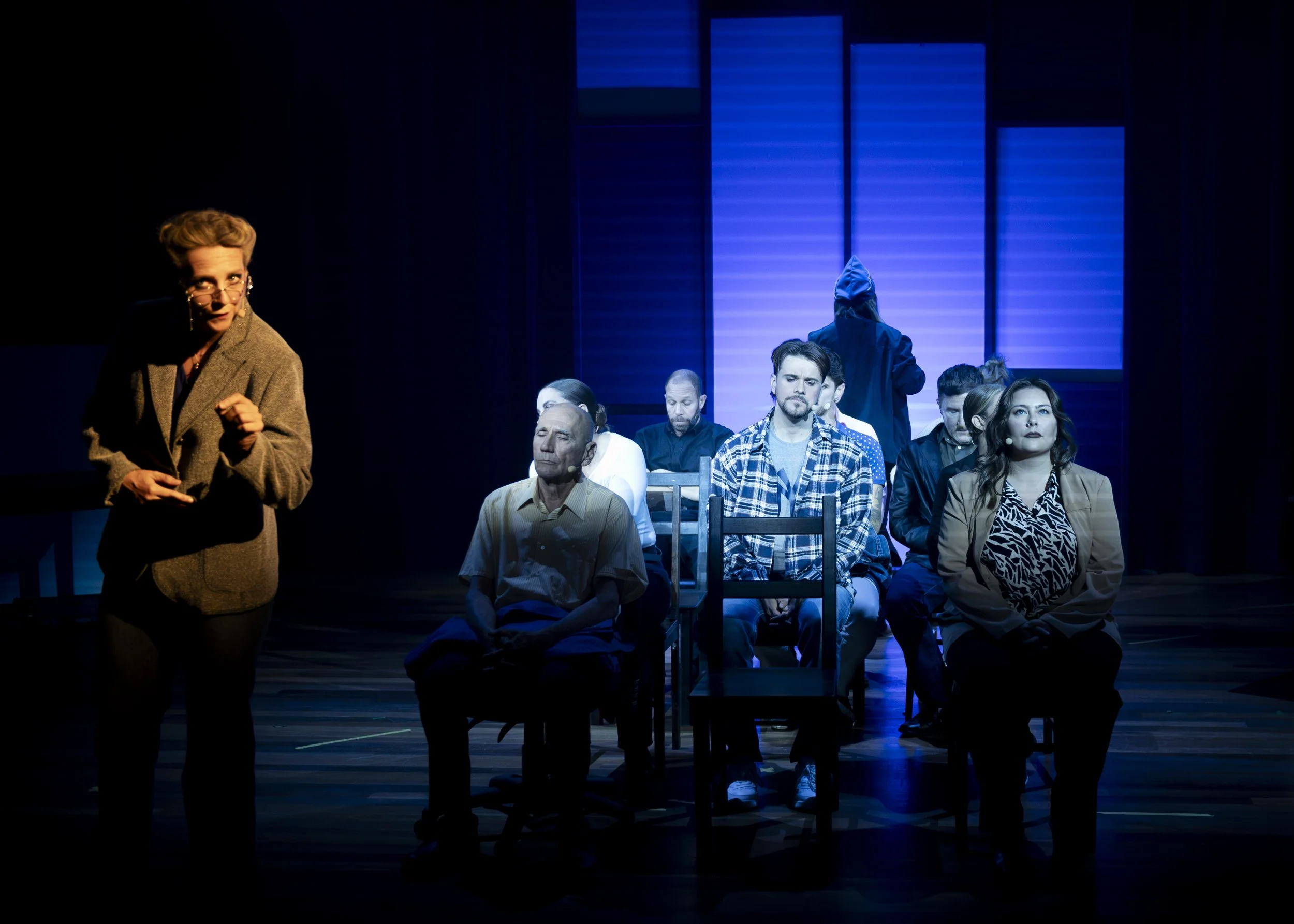 Stage scene with multiple actors seated in chairs, one woman standing on the left, performing or speaking. The background is lit with blue lights, suggesting a theatrical performance.