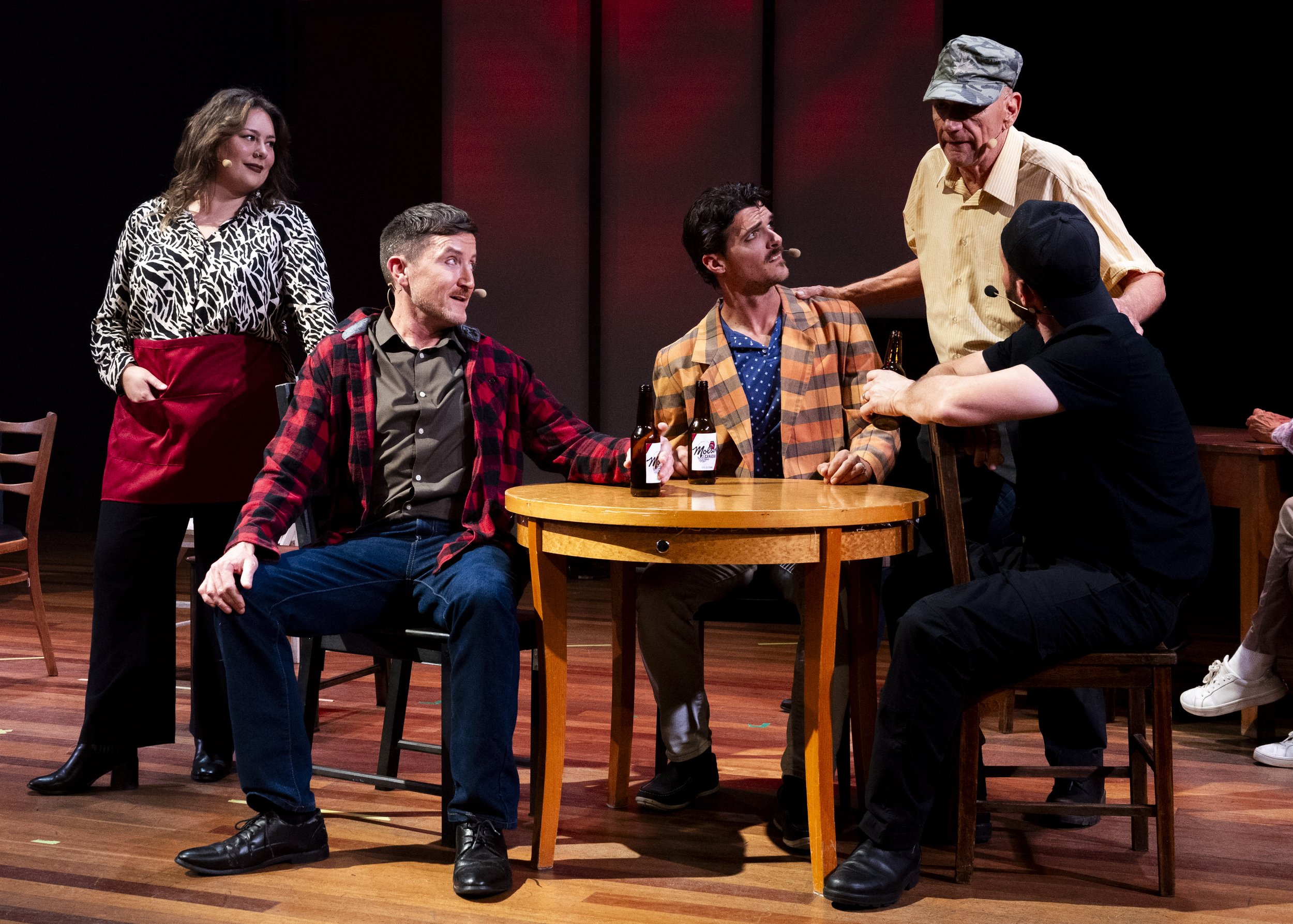 Six people are performing on a stage, with a table and chairs. One woman is standing on the left, wearing a zebra print blouse and red apron, looking at the others. A man in a red plaid shirt is sitting, holding a beer, and looking up at another man.