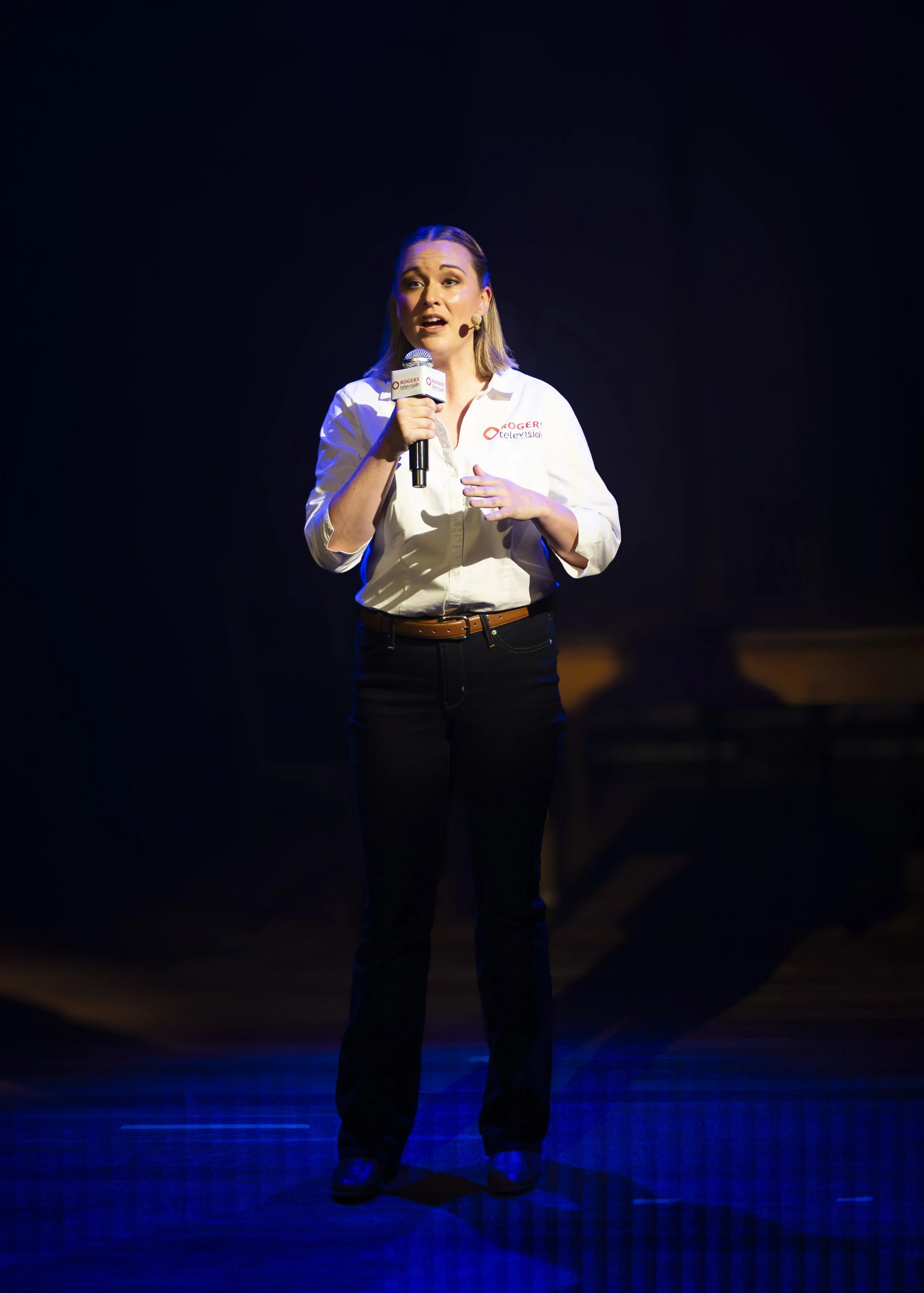 A woman speaking on stage, holding a microphone, wearing a white shirt with a Rogers logo, against a dark background with blue stage lighting.