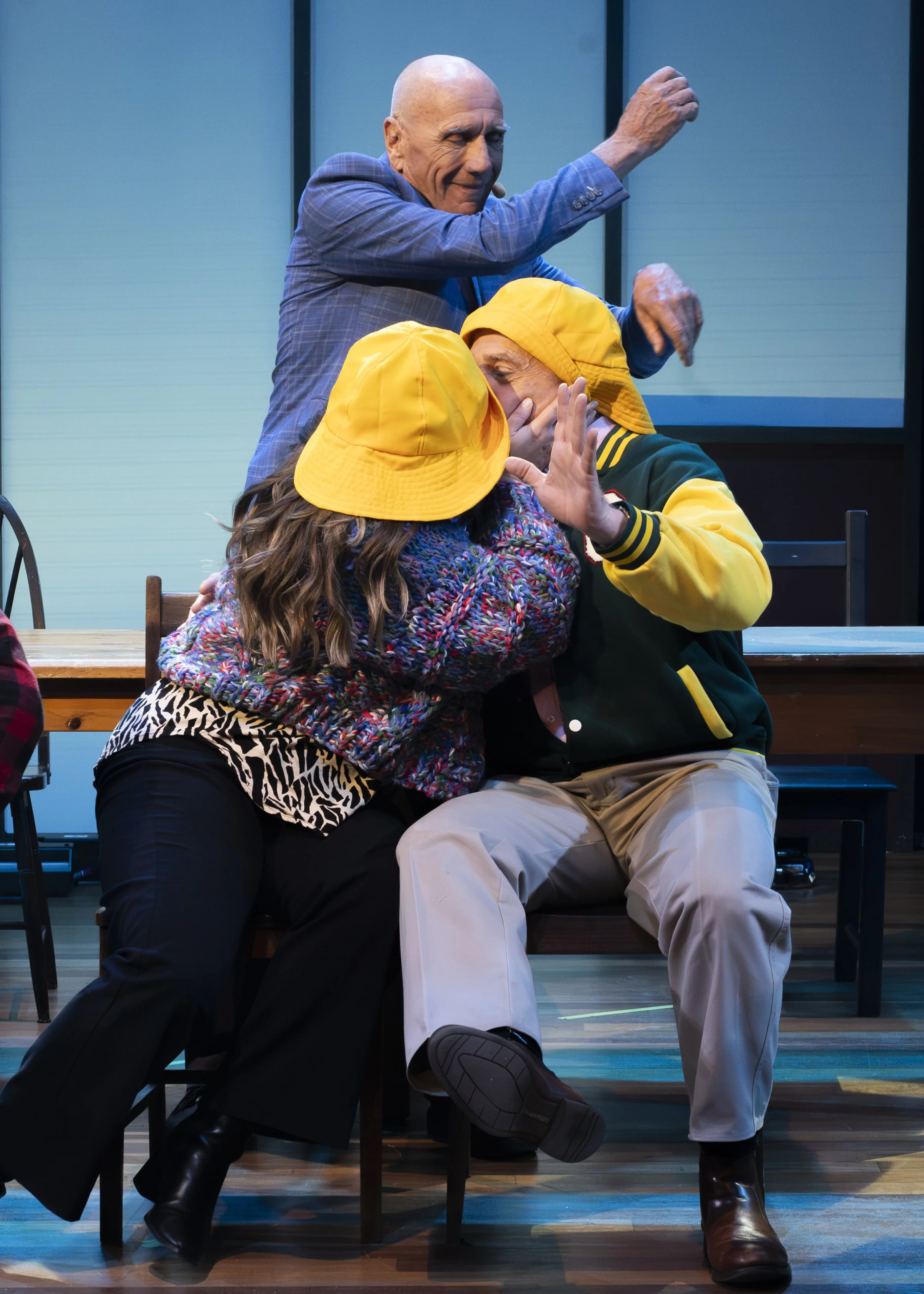 Three actors performing on stage, with one man standing behind two seated individuals, all wearing colorful costumes and expressive in their gestures.