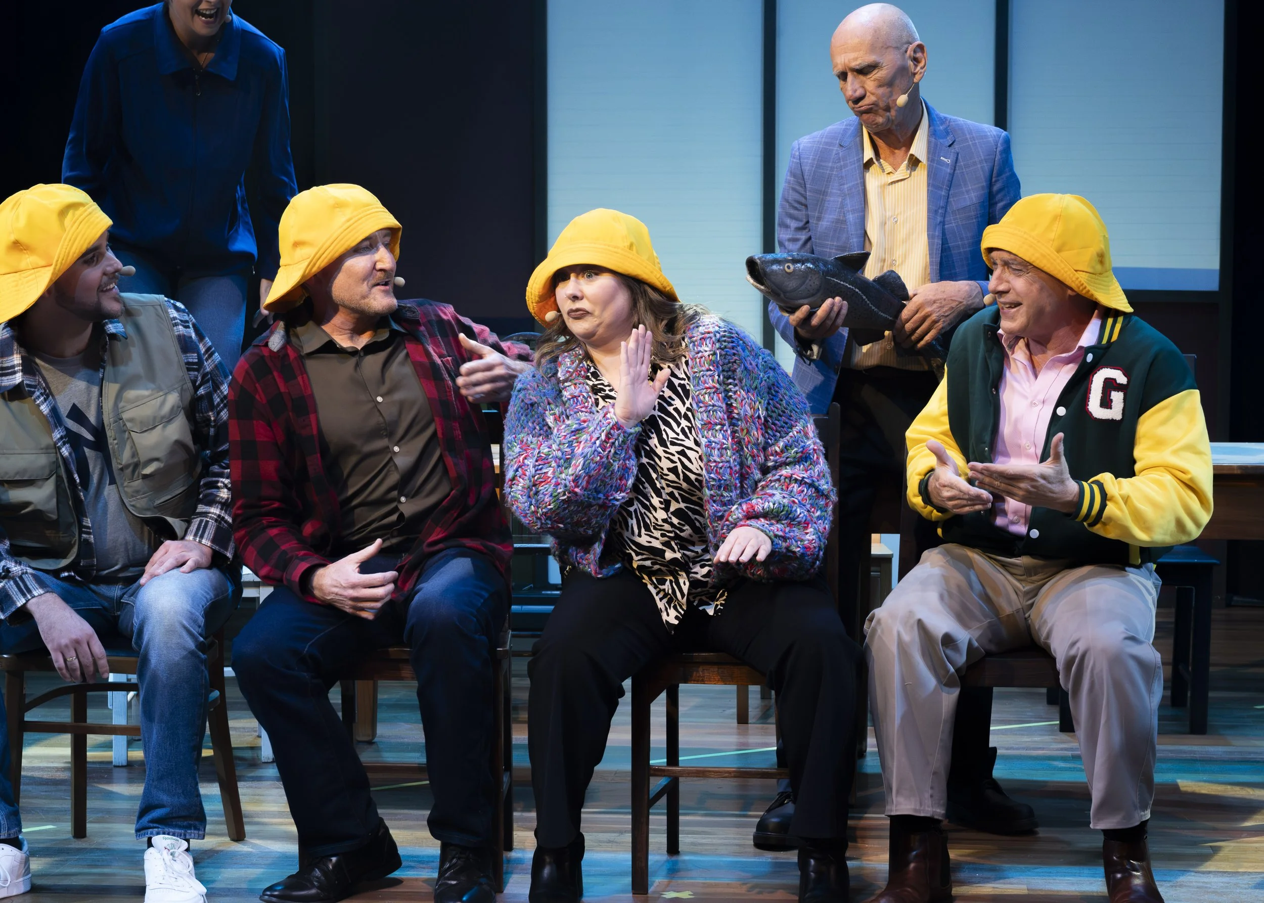 A theatrical performance with actors dressed as fishermen, some wearing yellow hats, and one holding a fish prop. They are seated and standing on stage, engaging in a comedic scene.