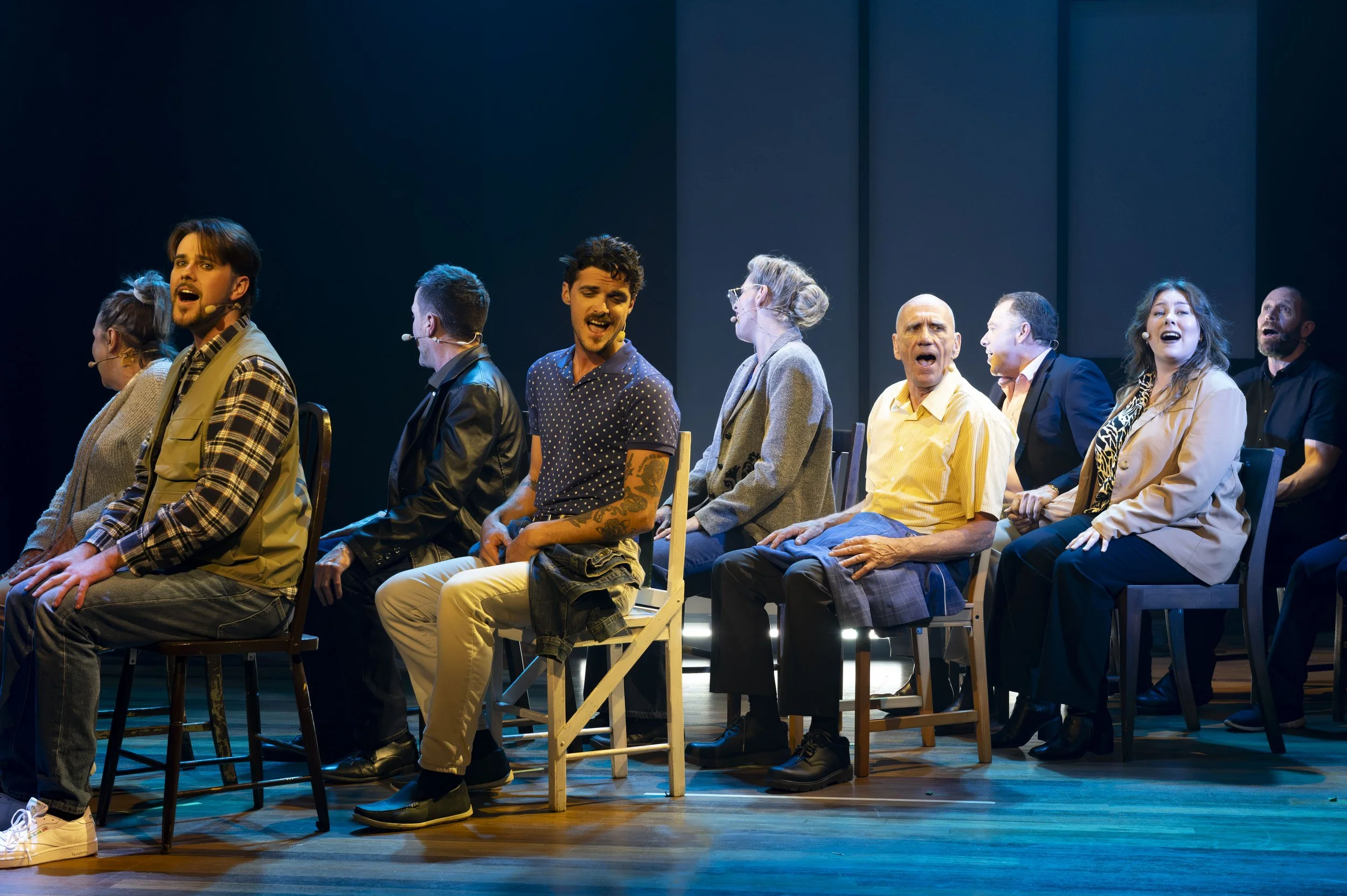 Actors sitting in a row performing on stage, singing or speaking, with plain dark background and stage lighting.