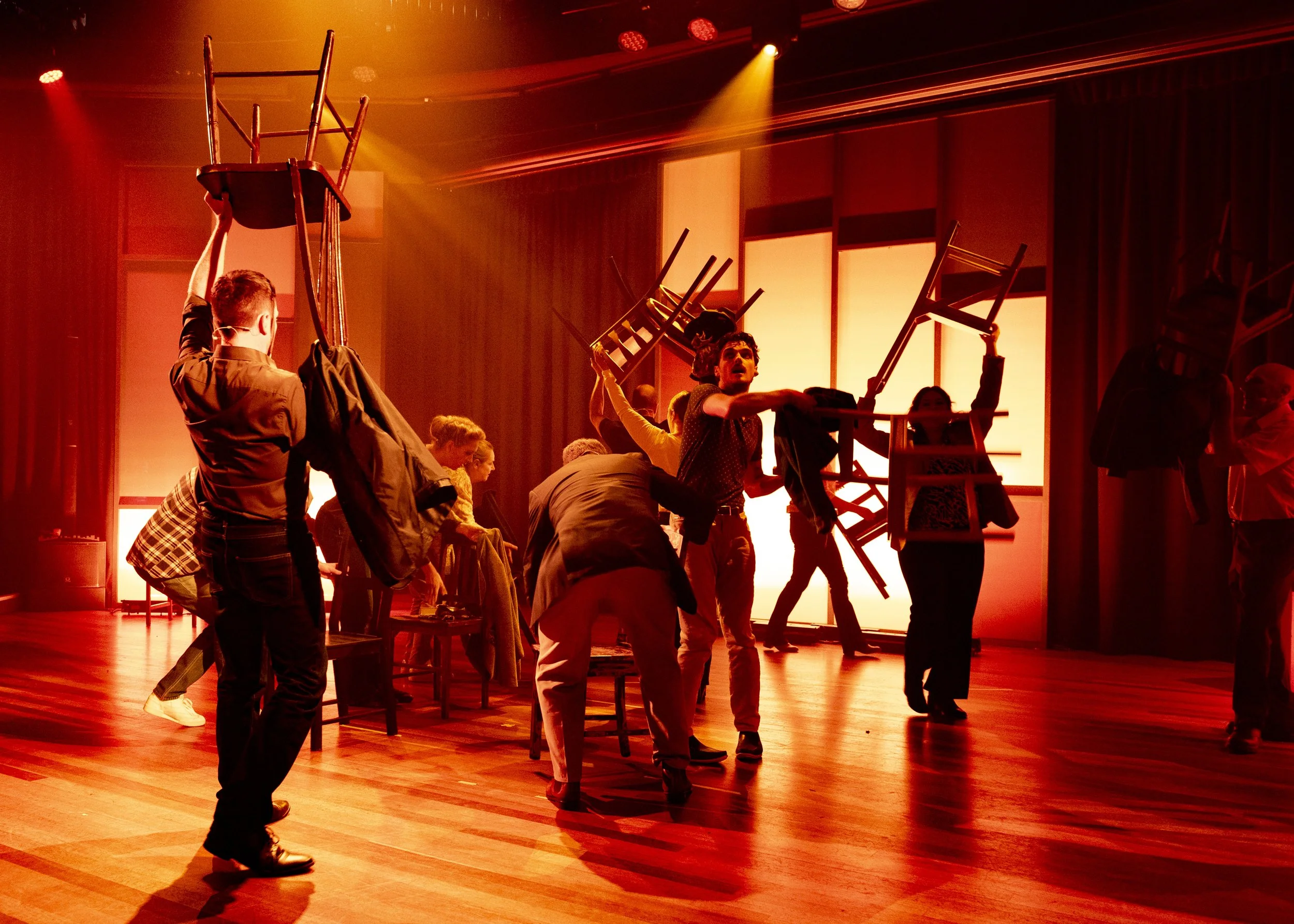 People on stage rehearsing with chairs during a performance, colorful stage lighting.