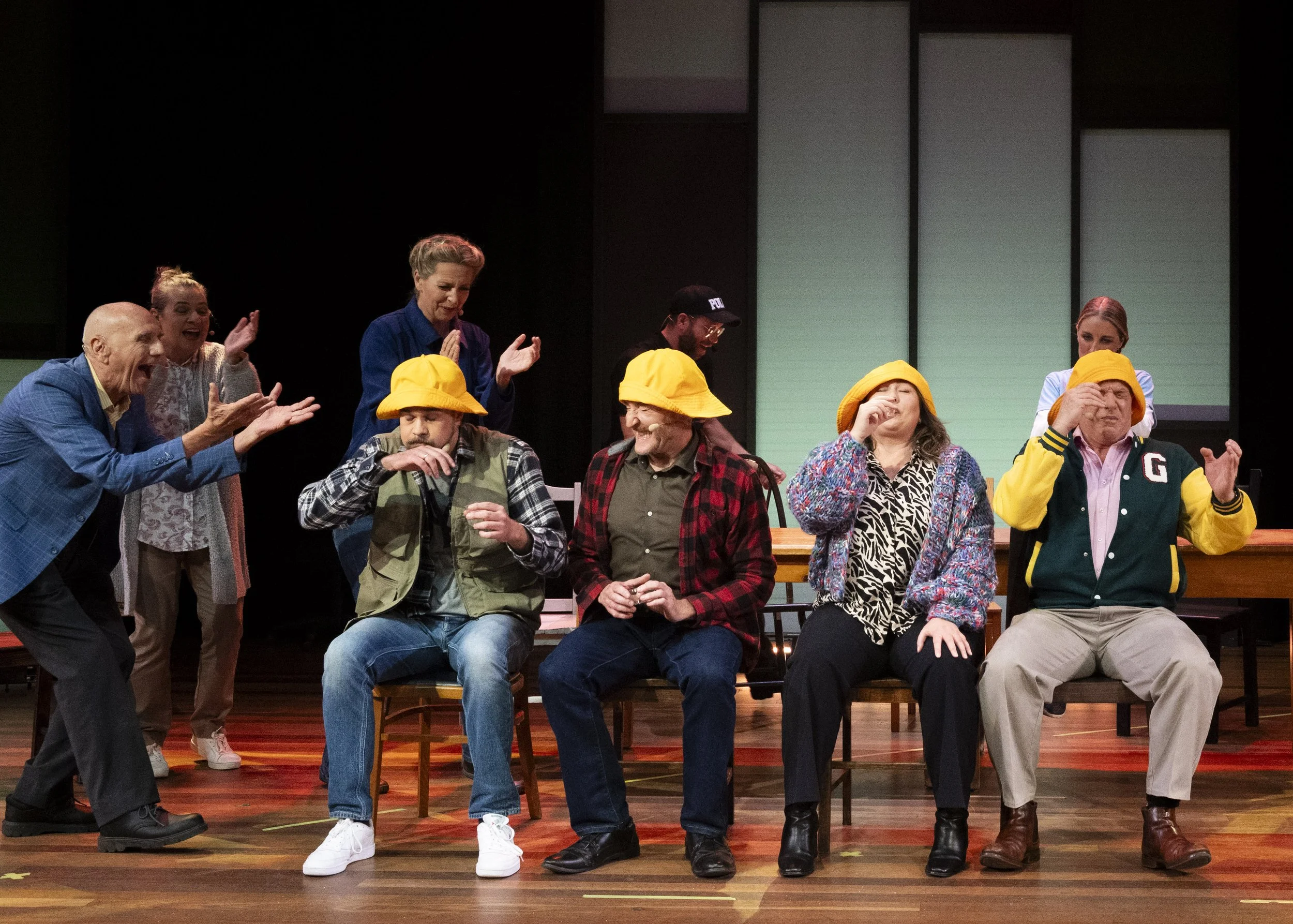 The image shows a stage with five seated people wearing yellow hats, with three individuals standing behind them. The seated individuals appear to be playing a game, possibly a blindfolded test, with one person in a green vest, another in a red plaid