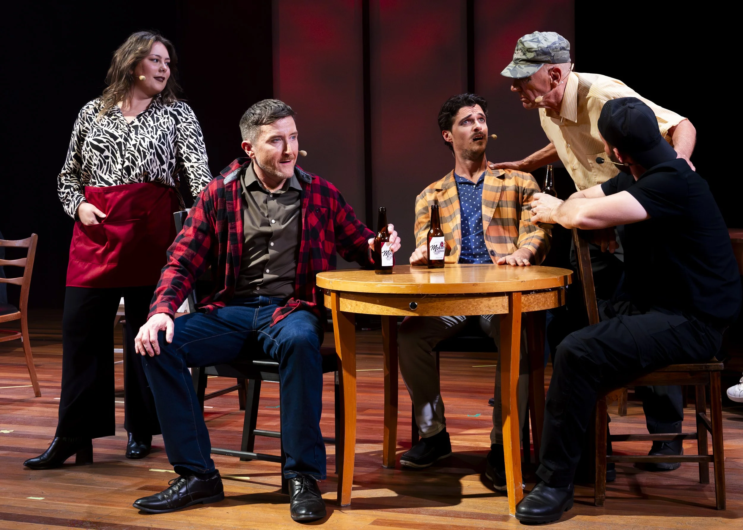 A theatrical stage scene with five actors, three seated around a round wooden table, two standing nearby. The actors are engaged in a dramatic or comedic moment, with some holding bottles of beer. The scene appears to be part of a play or performance
