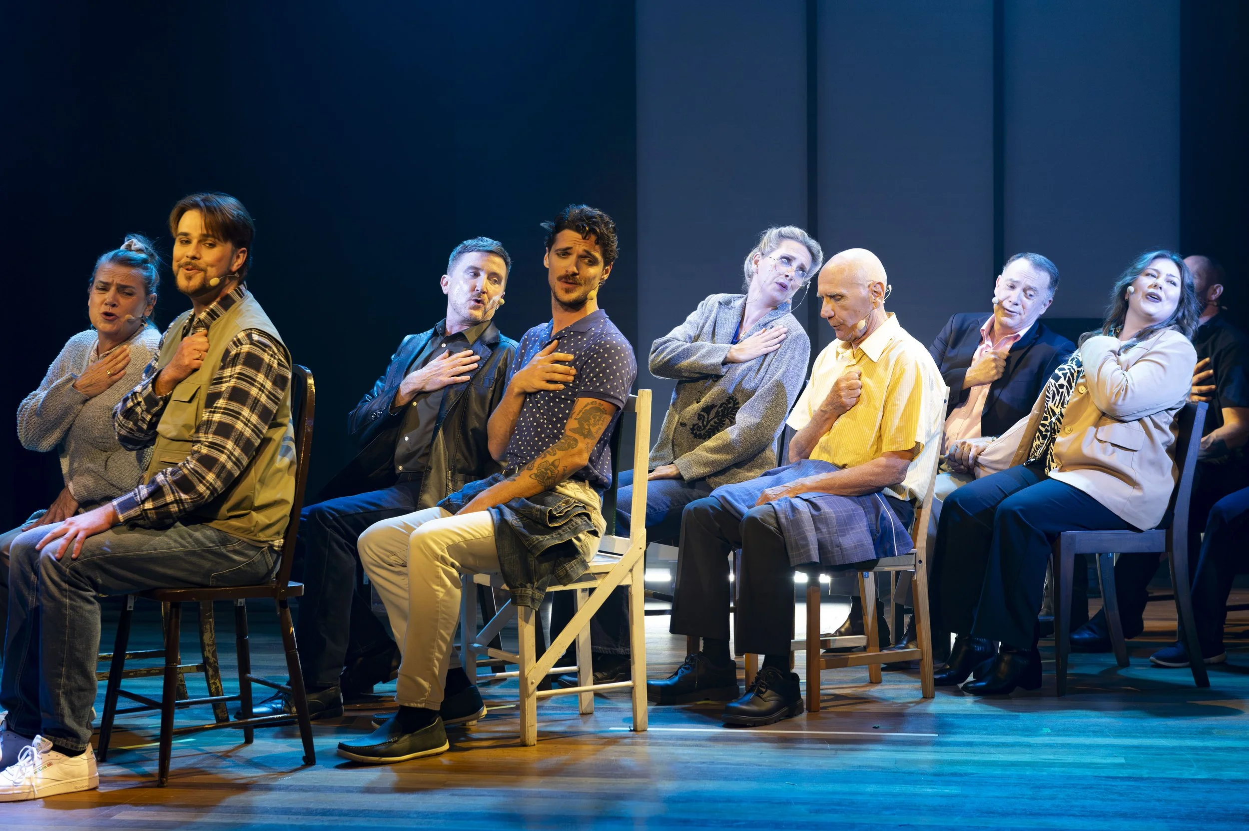 A group of diverse people sitting on chairs on stage, singing with their right hands over their hearts, during a theatrical performance.