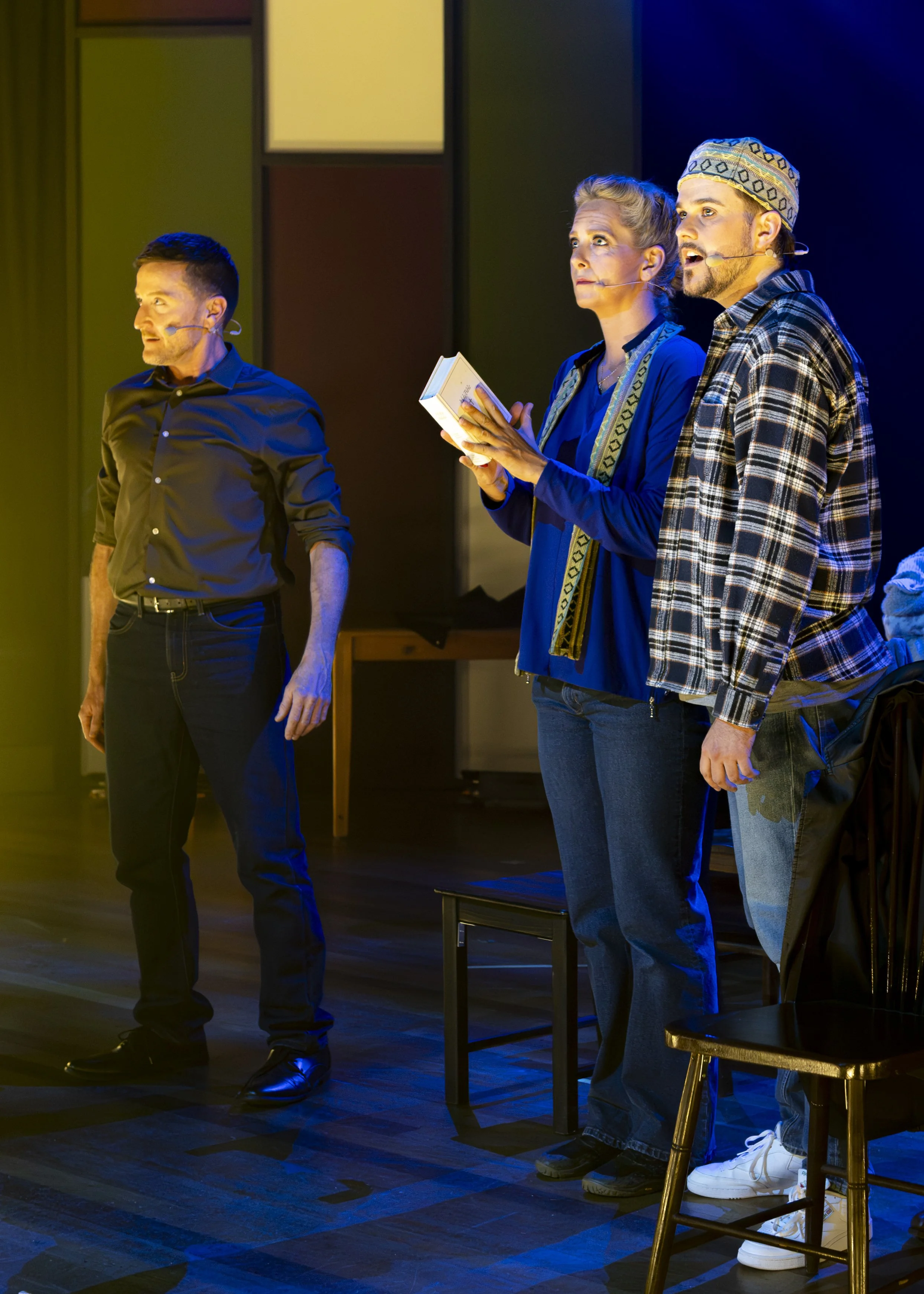 Three actors on stage during a theatrical performance, with microphones on their faces, lights illuminating them against a dark background, one woman holding a book, all appearing engaged in a scene.