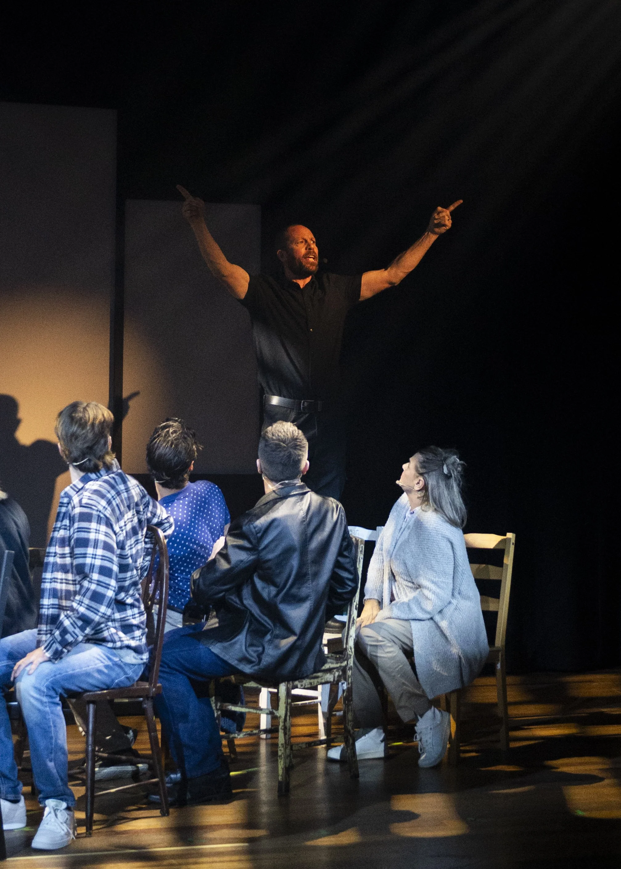 A man standing on a stage in front of a seated group, gesturing upward with both arms while speaking passionately.