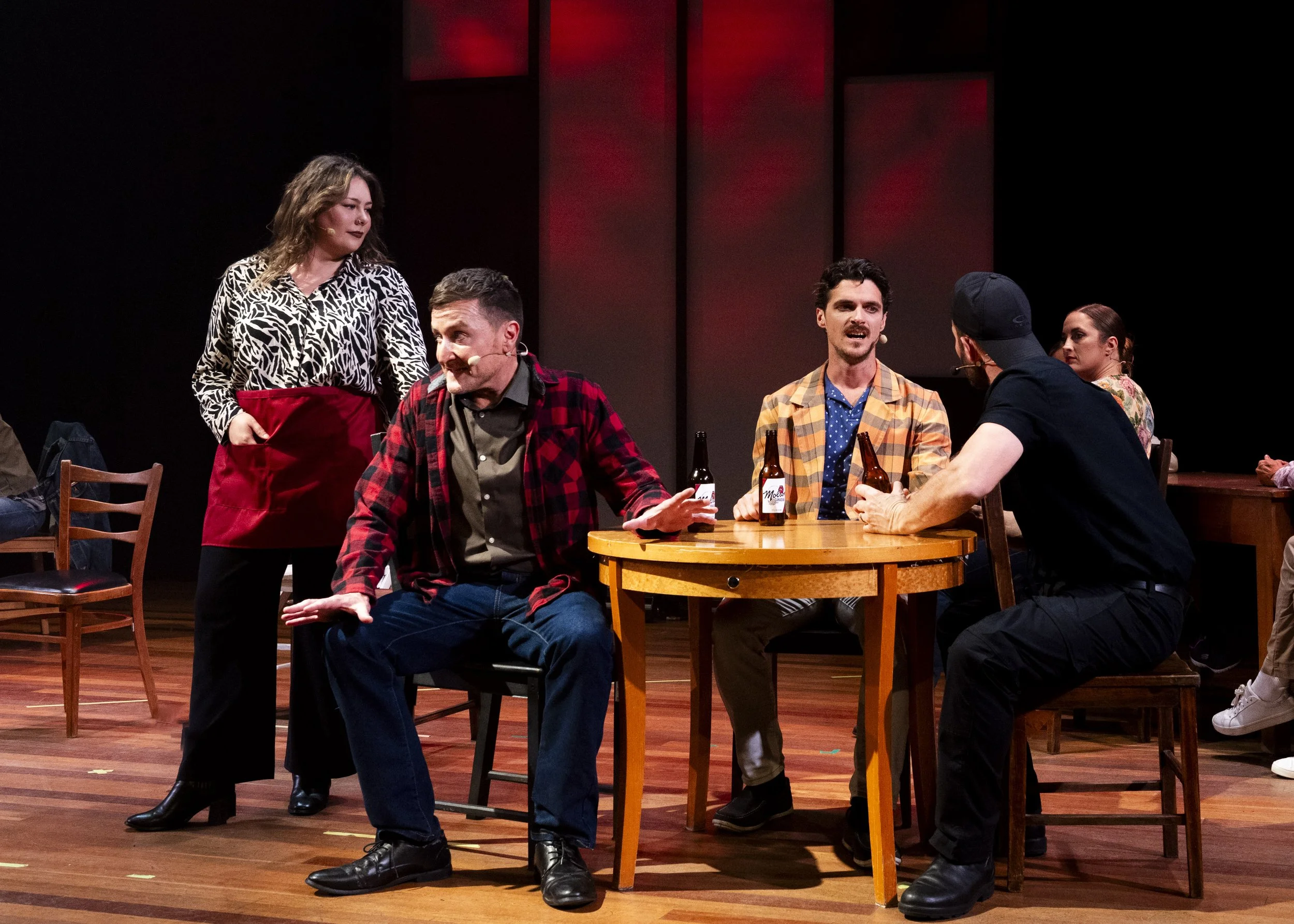 A theatrical play scene with five actors; three men and two women, engaged in a conversation at a round wooden table with three beer bottles. One man is speaking, seated on a chair, wearing a plaid jacket. The woman standing next to him has her hands