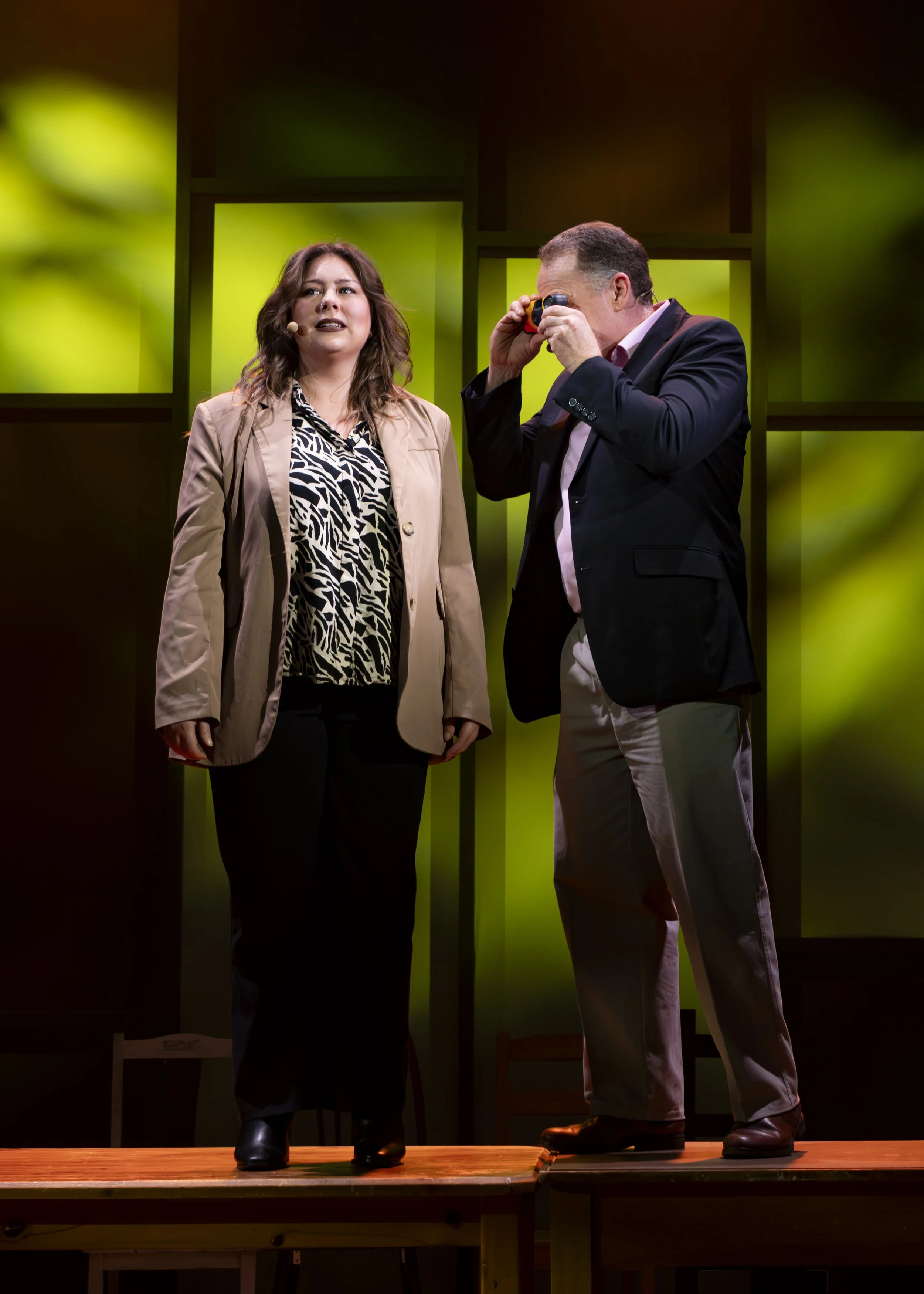 A woman with long brown hair wearing a beige blazer and animal print blouse standing on stage next to a man with short hair in a dark blazer holding a small device to his eye, with a background of green lit panels.