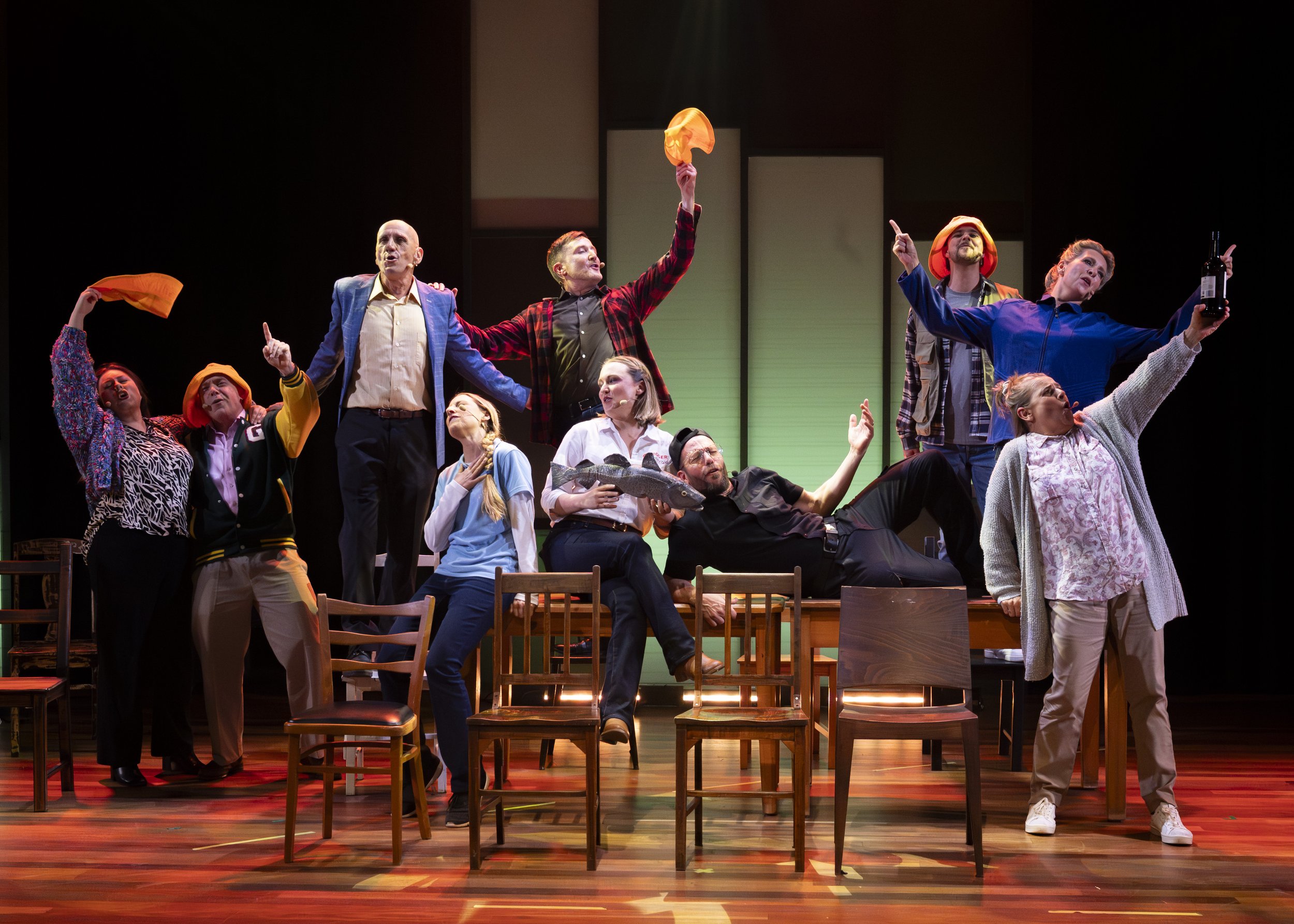 A theatrical stage scene with a group of actors performing. Some actors are standing, others are seated or lying, and they are dressed in colorful costumes. The scene is lively, with actors gesturing and expressing enthusiasm.