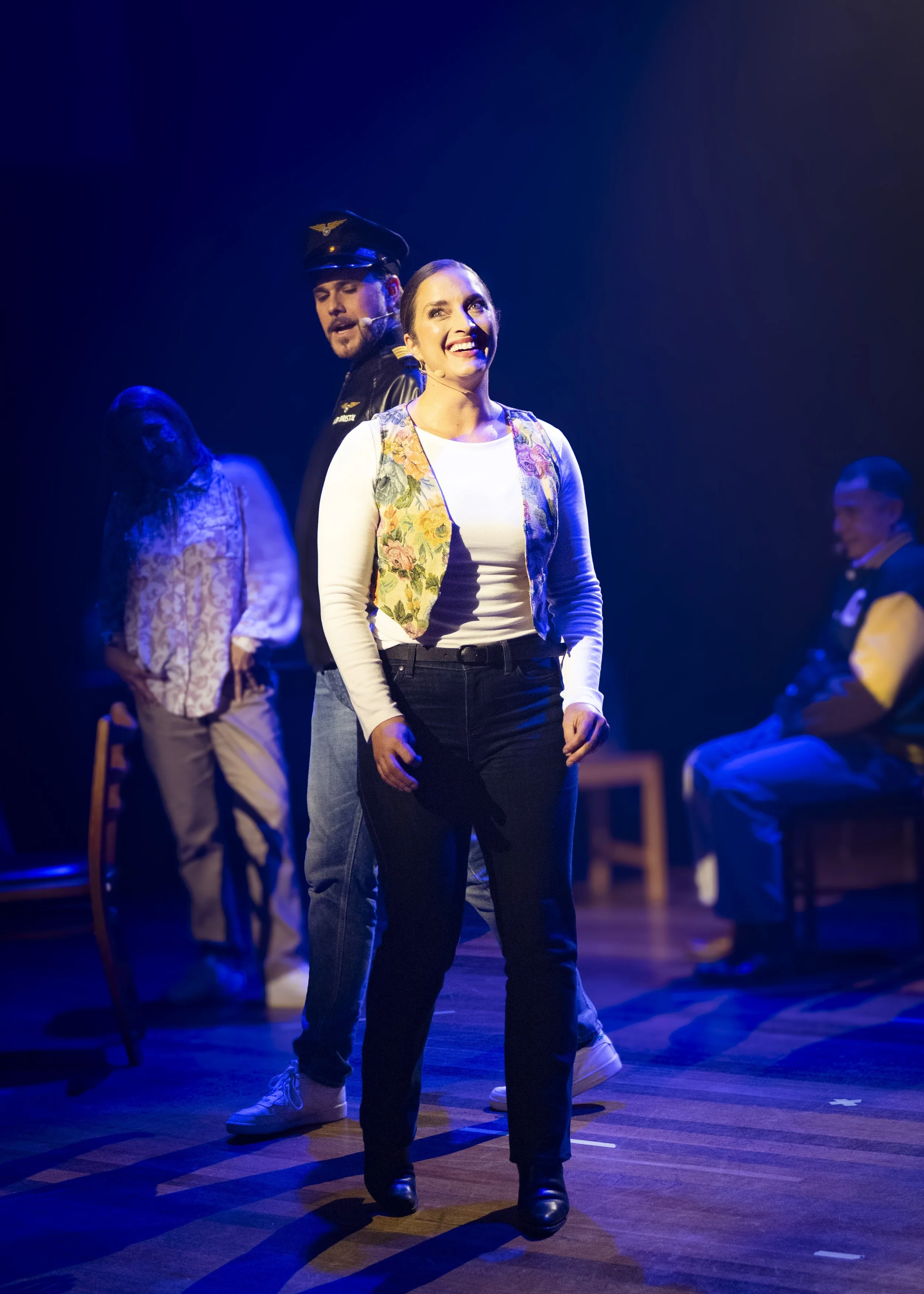 People performing on stage with dark background, woman smiling at center, three other performers in the background.