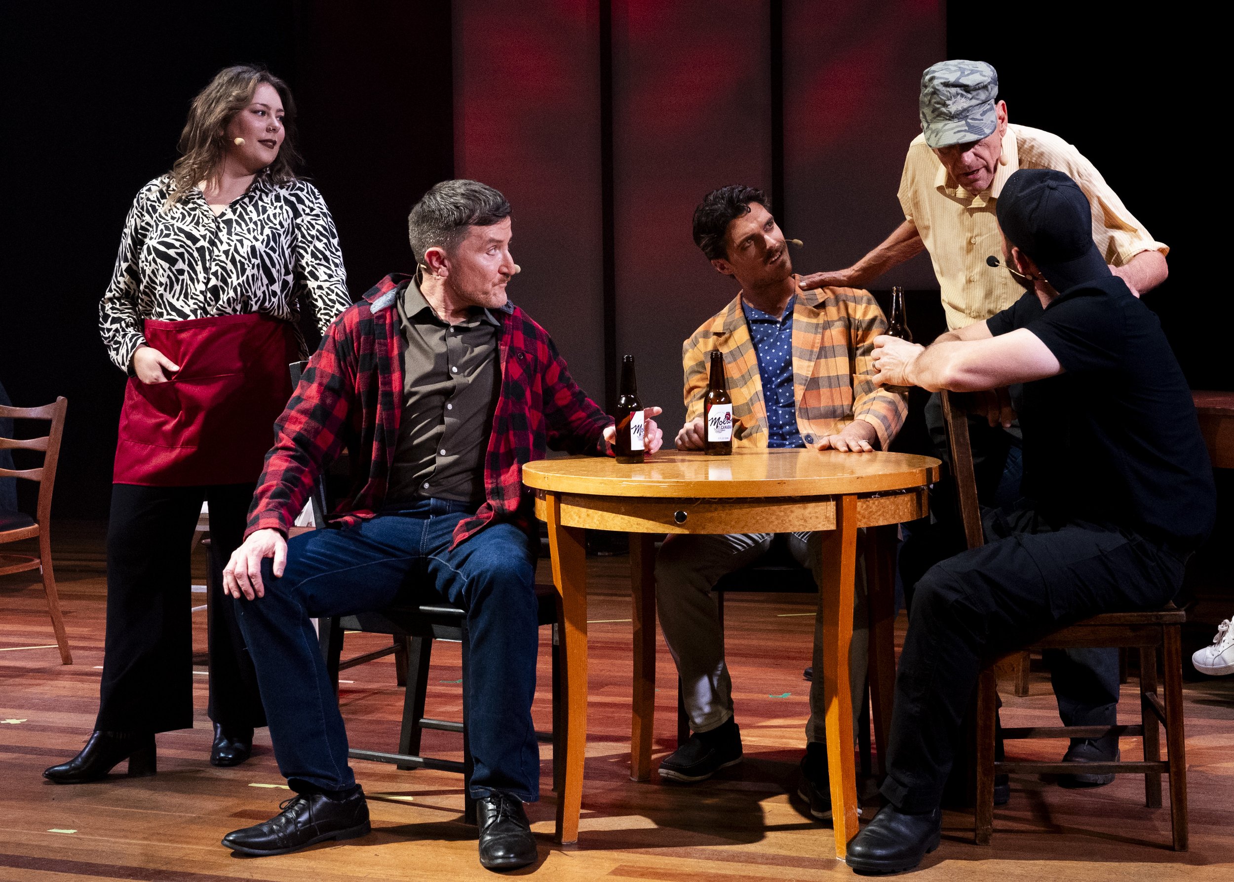A theatrical scene with five actors around a wooden table holding beer bottles. One man in a military cap is talking to a man in an orange checkered shirt, while a woman standing nearby wears a black and white patterned shirt and red apron, observing