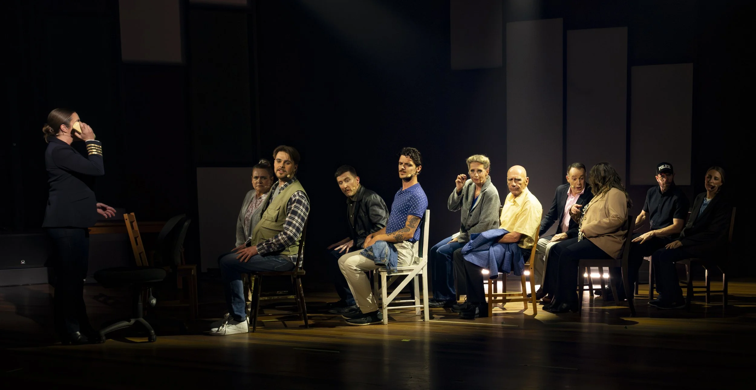 The image shows a group of people sitting in a row on stage, facing the woman on the left, who is standing and holding a phone to her ear, dressed in a pilot or naval officer uniform. The stage is dimly lit with a dark background, and the people appe