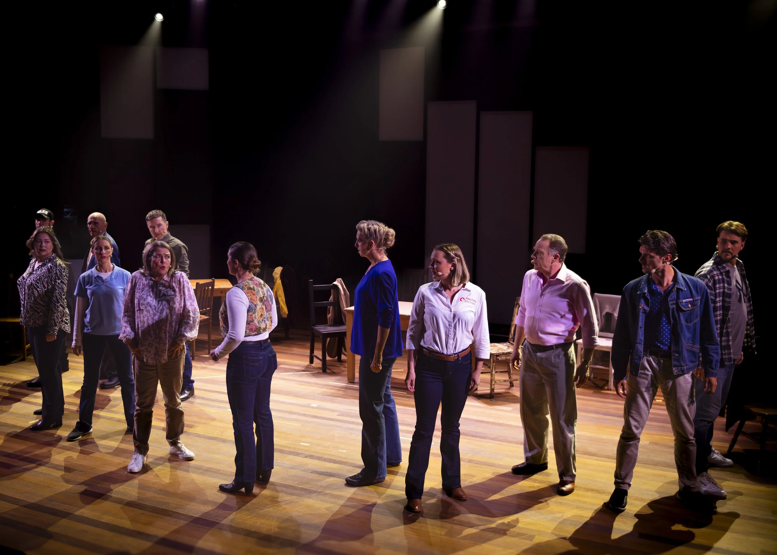 A group of people standing in a line on a stage with wooden flooring, some chairs and tables in the background, and dramatic lighting from above.