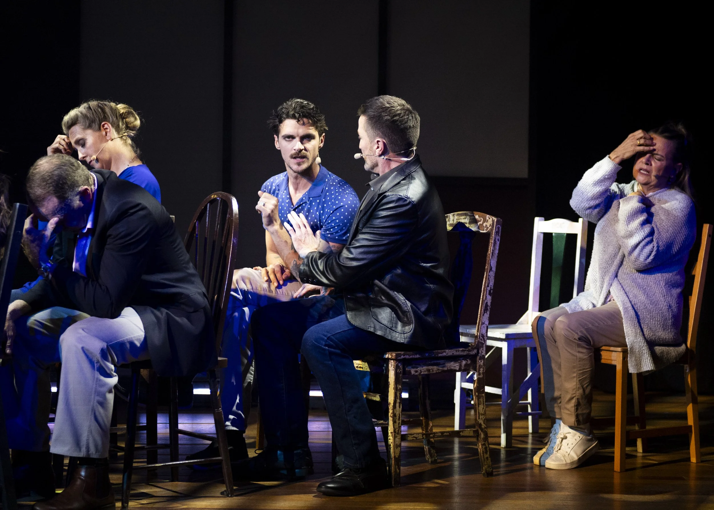 People sitting in a row on chairs on a stage, appearing to be in emotional distress or deep thought during a theatrical performance.