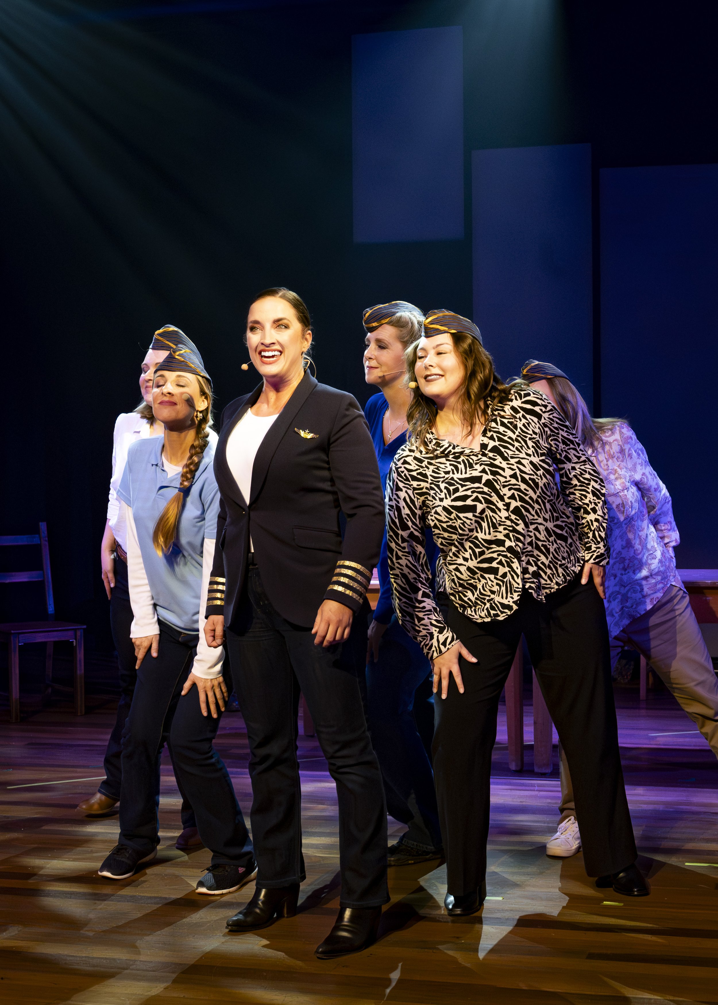 A group of women on stage, dressed in pilot or flight attendant uniforms and casual clothing, performing a musical or theatrical scene with some women wearing hats, under stage lighting.