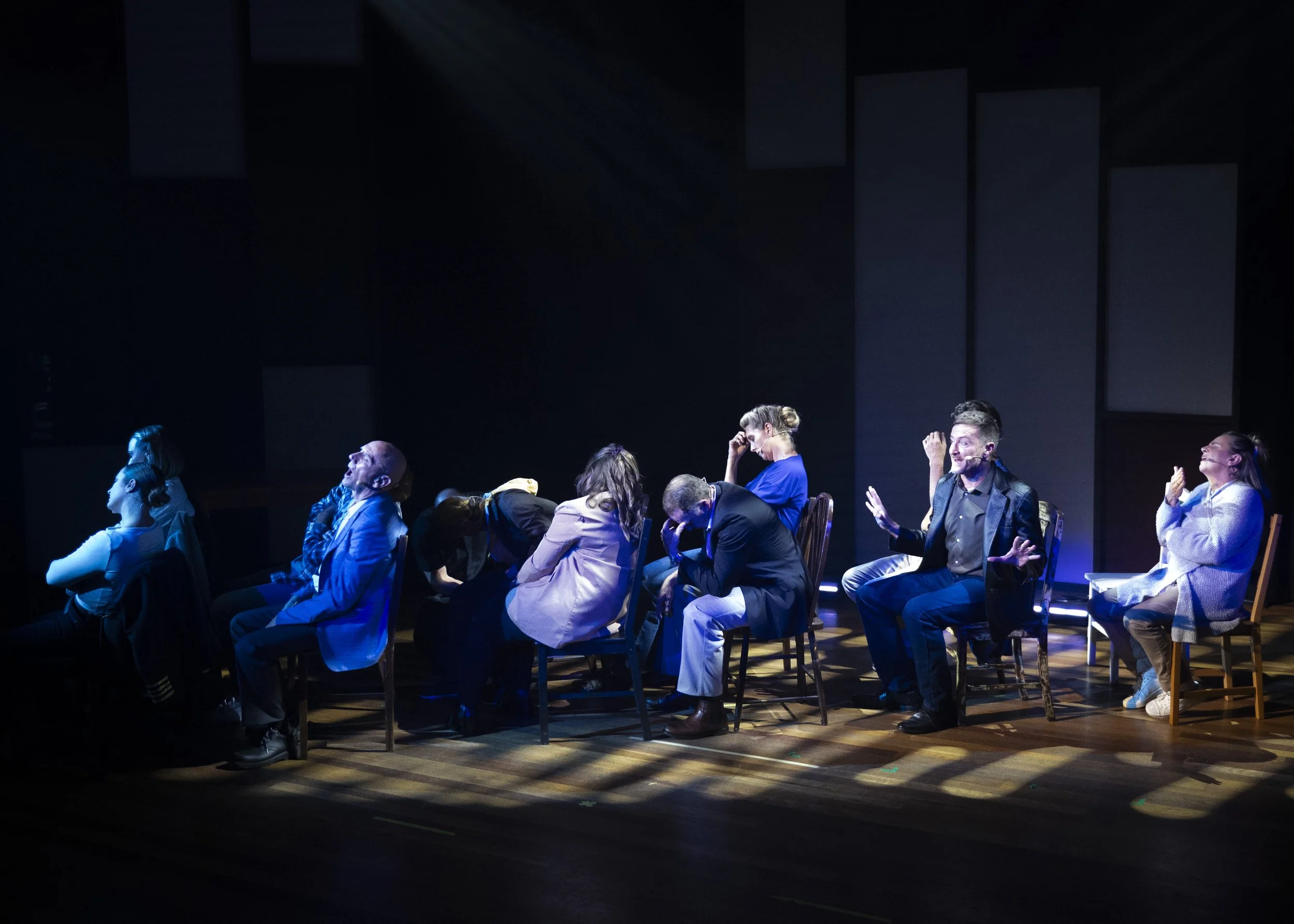 Group of people sitting on chairs on a stage, some appear to be laughing or crying, in a theatrical lighting setting.