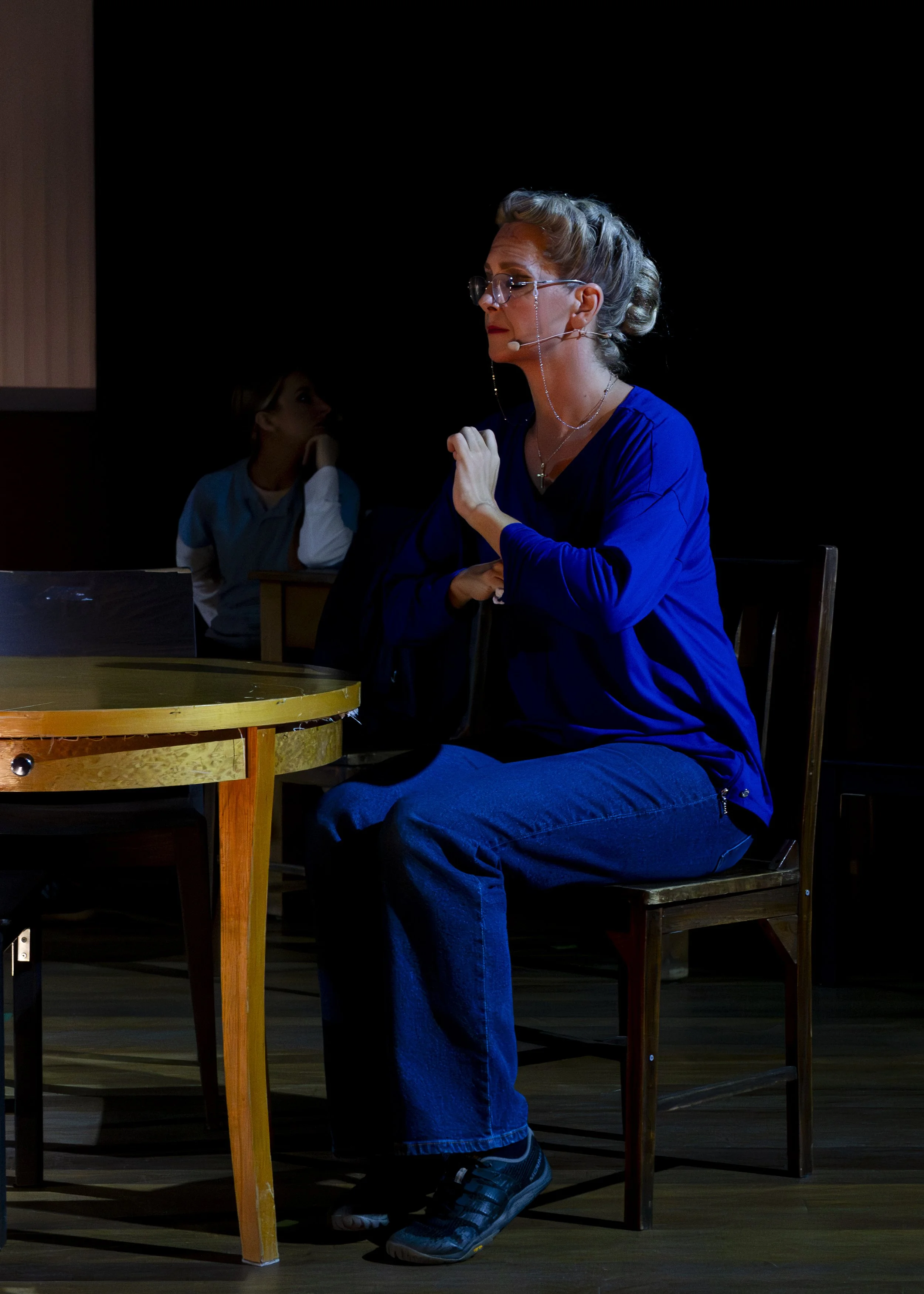 A woman with glasses and a headset, wearing a blue top and jeans, sitting in a chair at a table on a dark stage, with a young girl in the background.