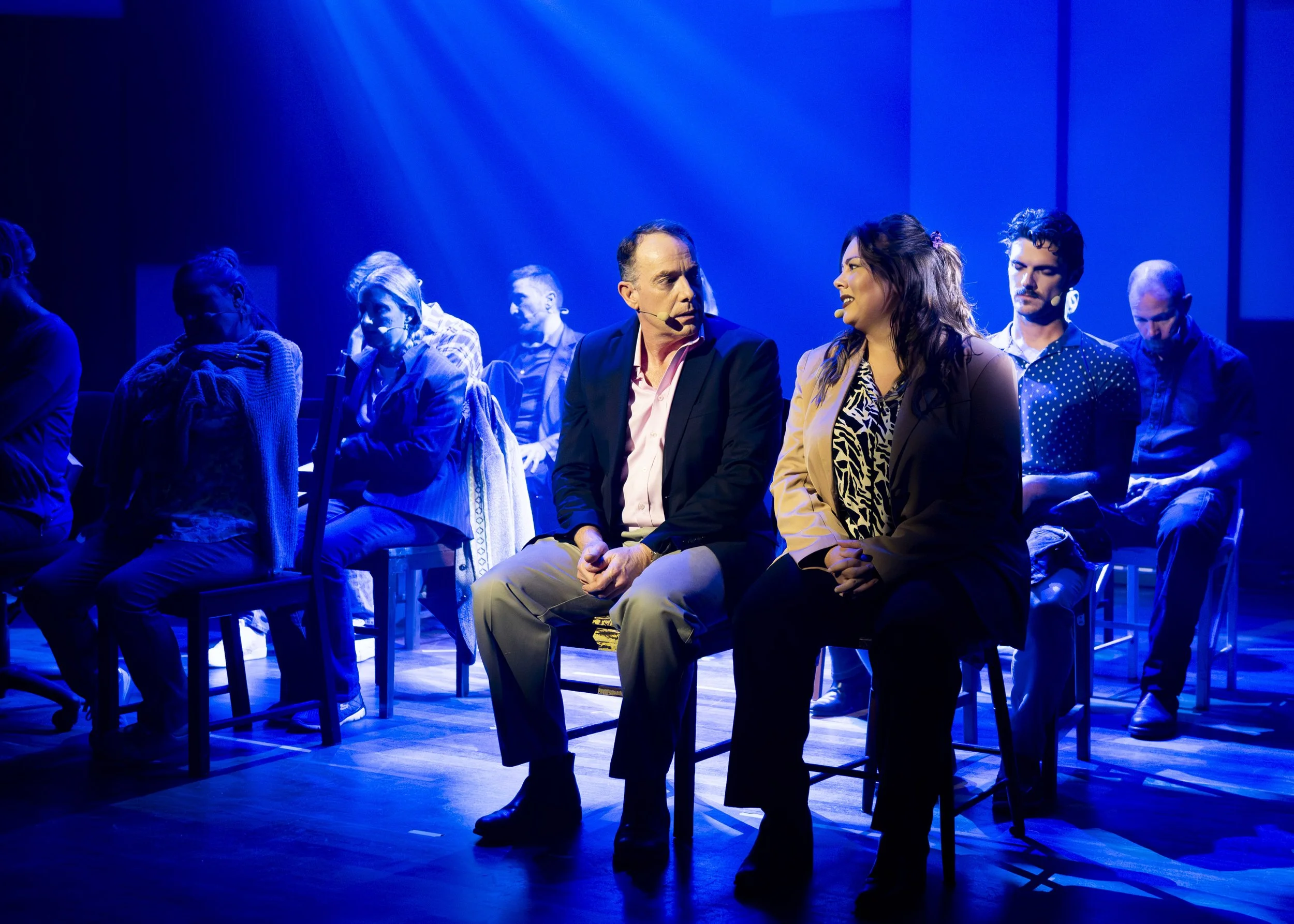 A group of people sitting in a row on stage under blue lighting, engaged in conversation, with two people in the foreground talking to each other.