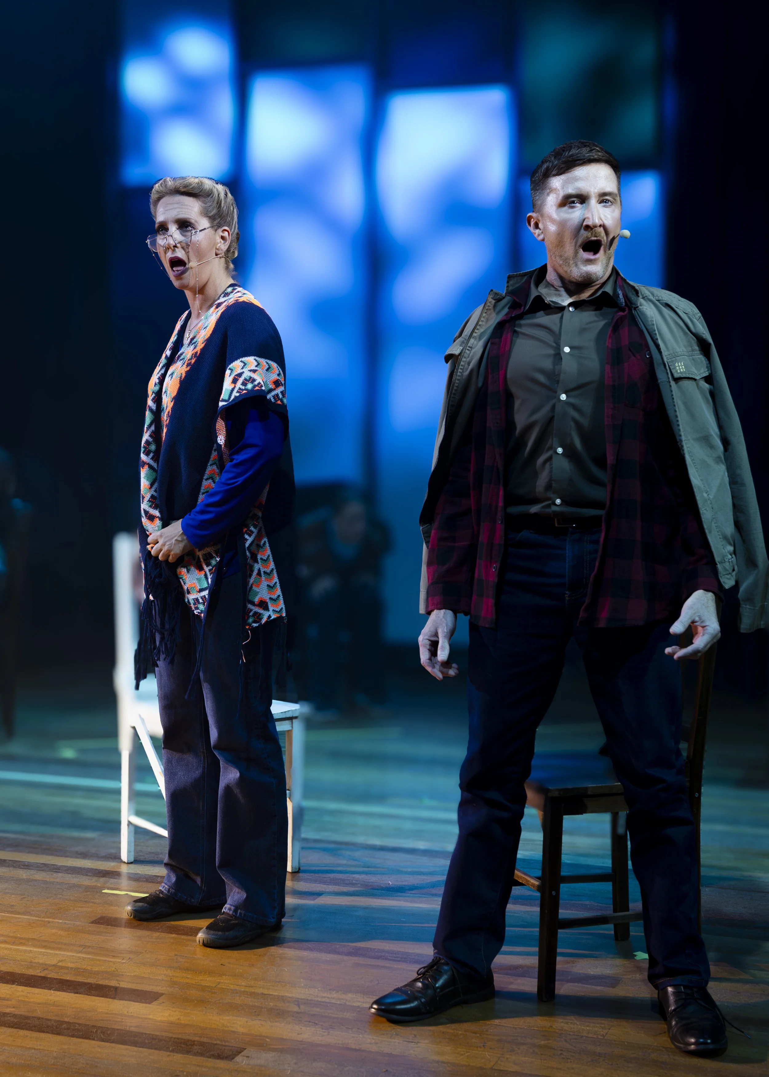 Two actors on stage, one woman and one man, singing with surprised expressions. The woman on the left is wearing glasses and a colorful, patterned shawl over a blue shirt and jeans. The man on the right is wearing a green jacket, a red plaid shirt, a
