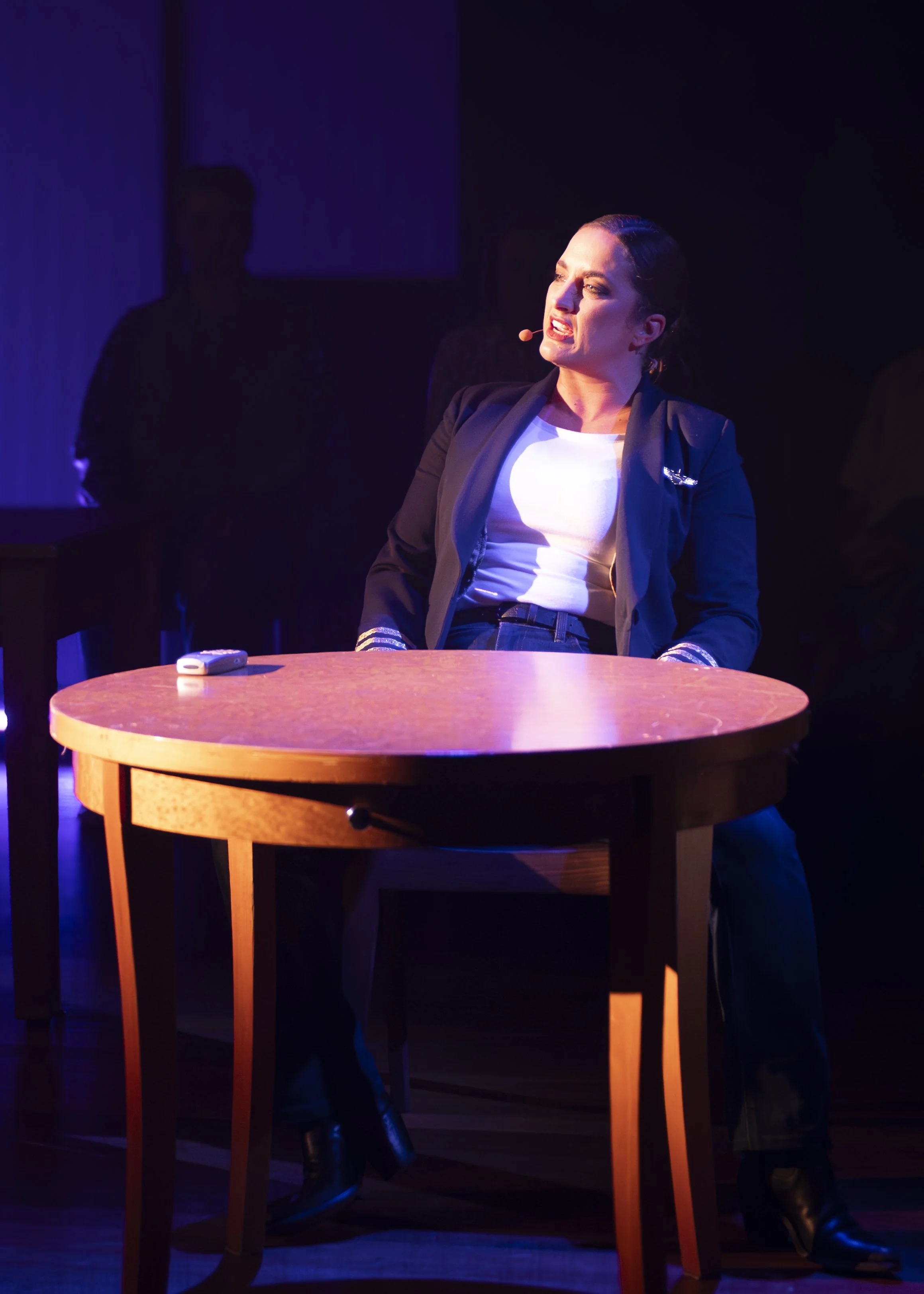 A woman performing on stage, sitting at a round wooden table, wearing a black blazer and white top, with a microphone headset, with dark background and purple lighting.