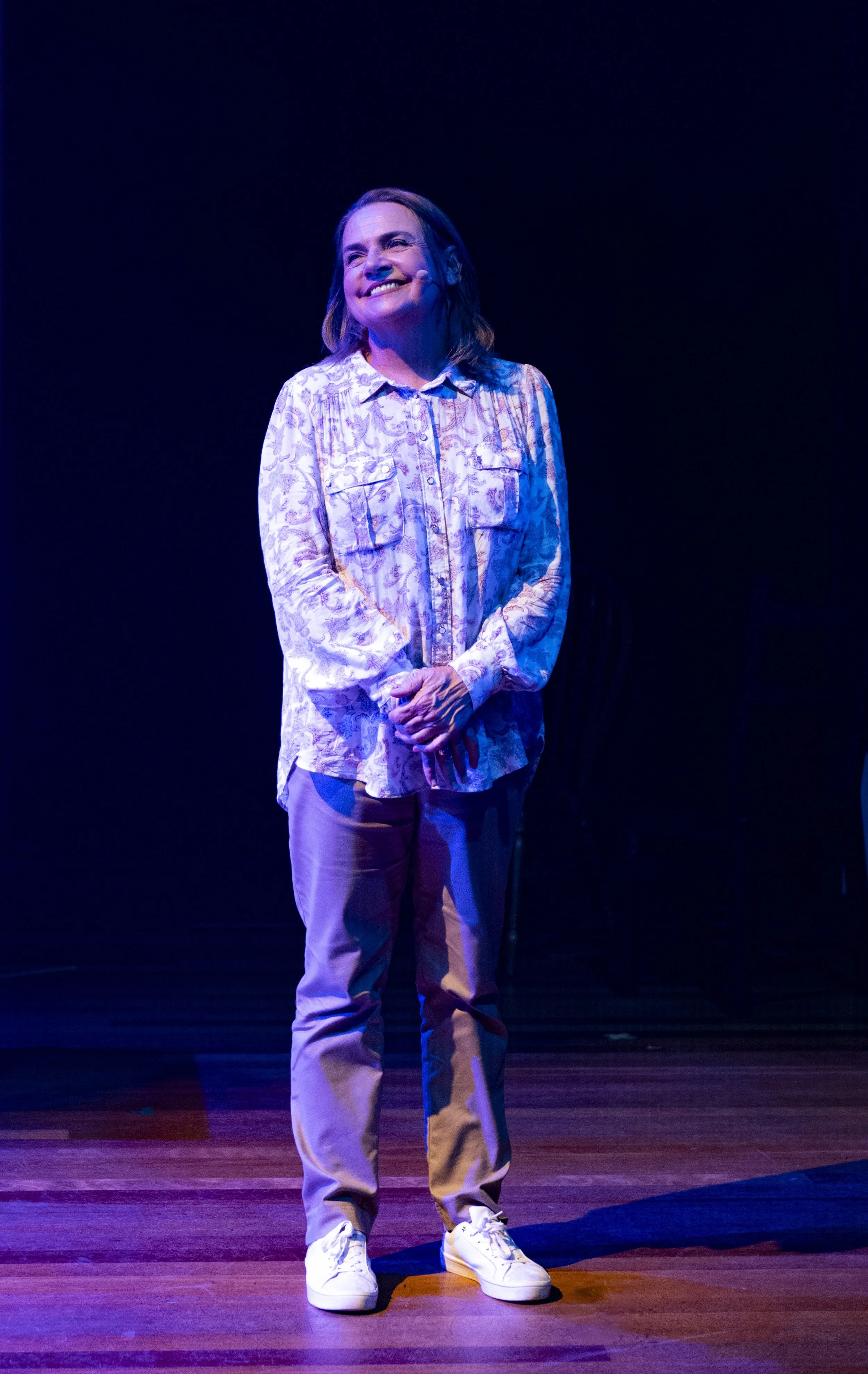 A woman standing on stage smiling, wearing a patterned long-sleeve shirt, khaki pants, and white sneakers, with stage lighting highlighting her against a dark background.
