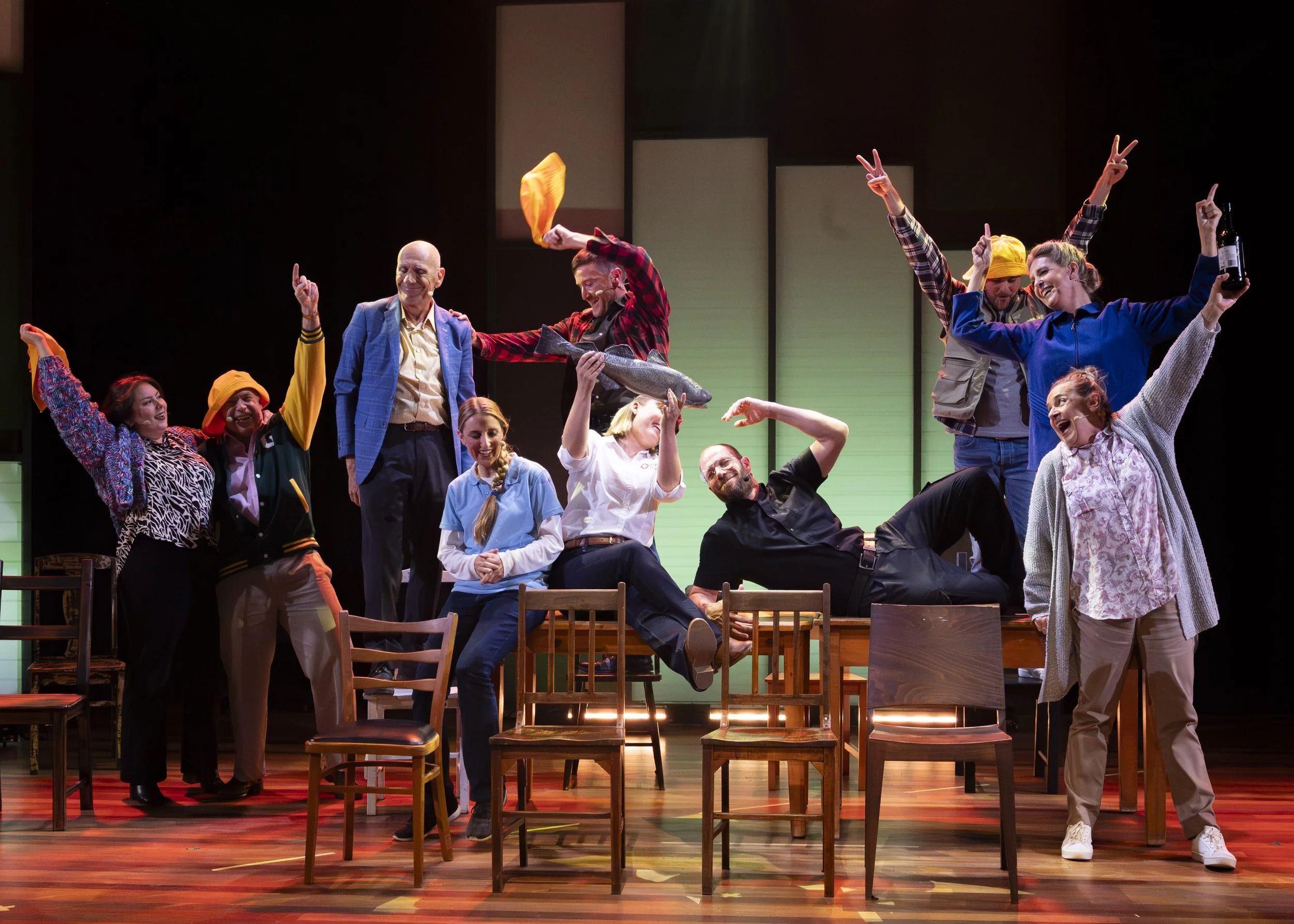 A group of actors on stage during a theater performance, with some standing and others sitting or lying on chairs, making expressive gestures and smiling, with a green backdrop.