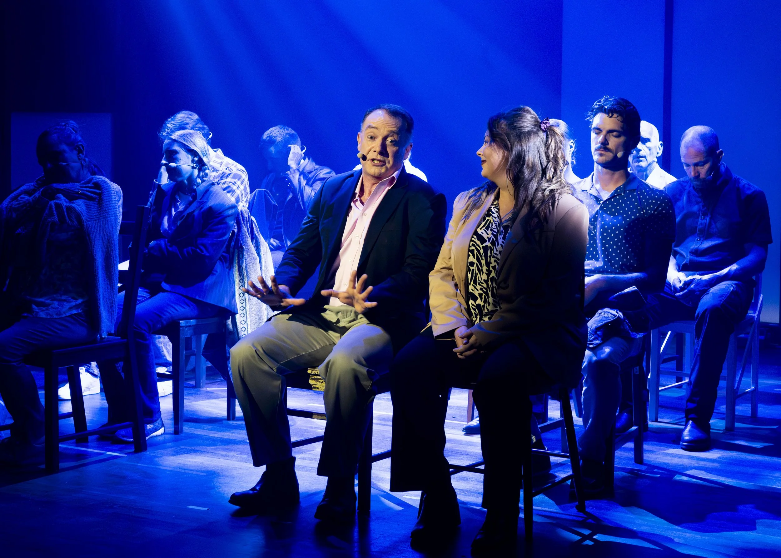 People attending a talk or performance on stage with blue lighting, seated on chairs in rows, engaged in conversation.