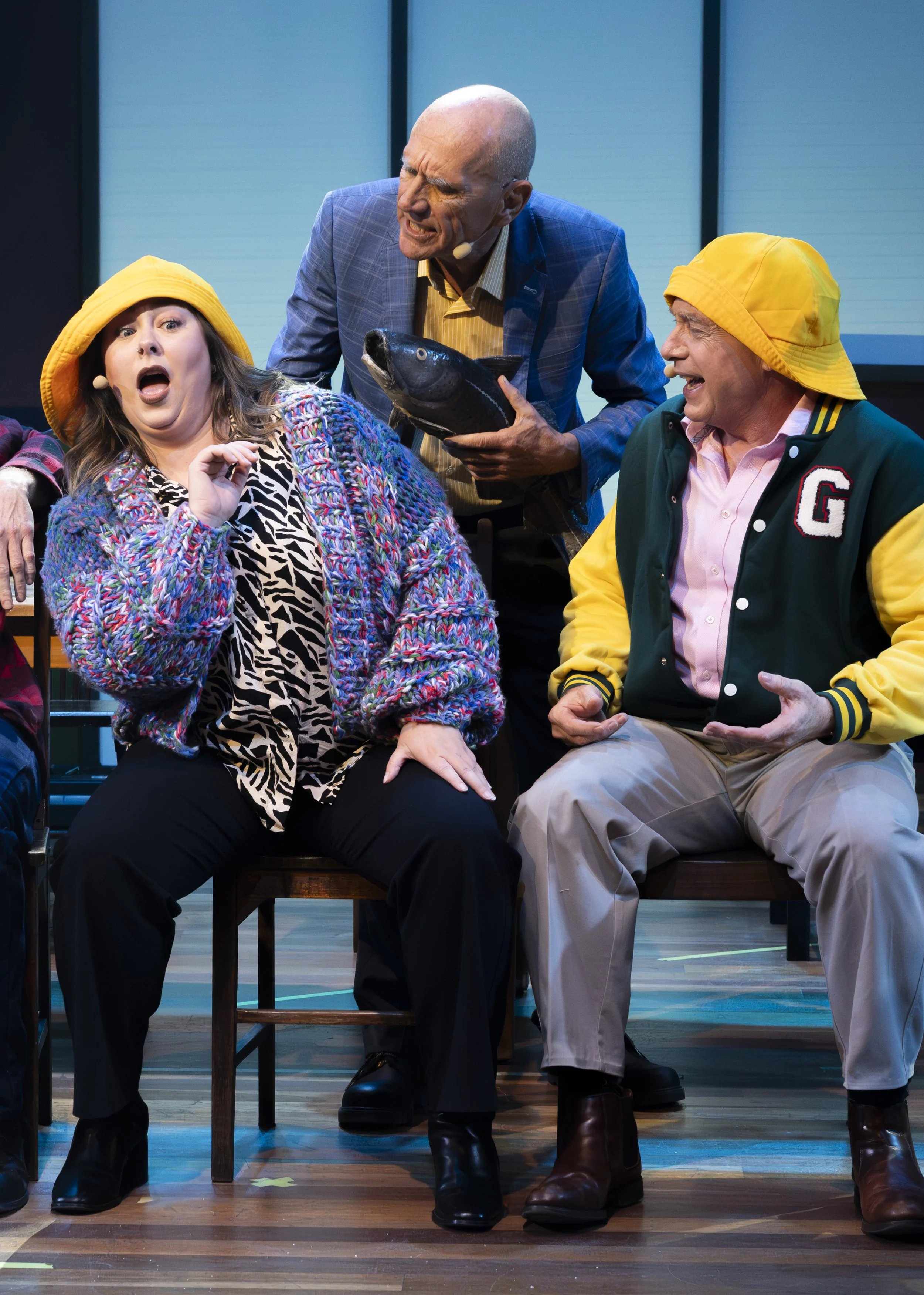 Three people on stage, two men and a woman, engaged in an animated scene with humorous expressions. The woman, seated, has a surprised look, wearing a yellow hat and colorful sweater. The man on the right, also seated, is smiling and wearing a yellow