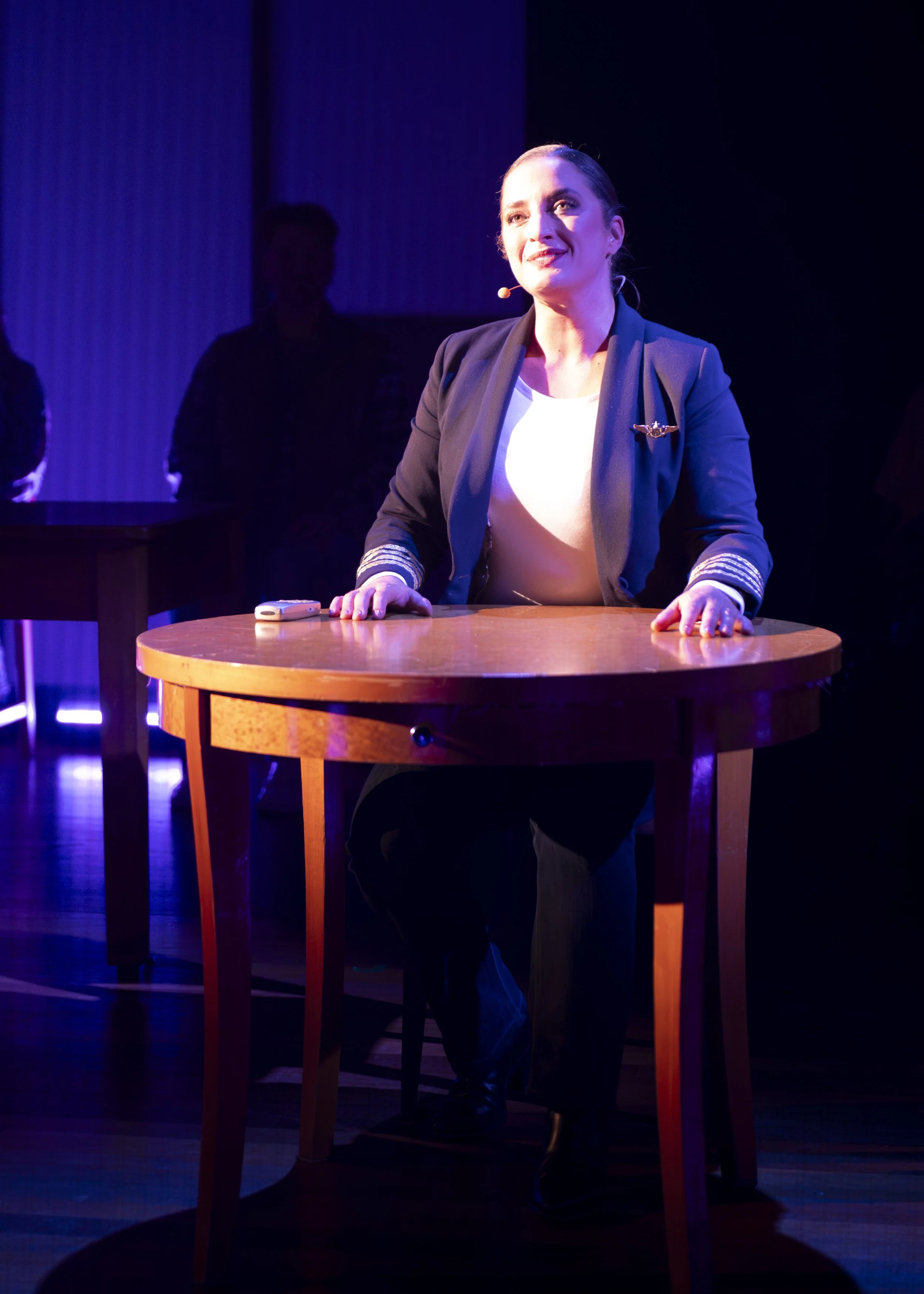 A woman with a microphone headset sitting at a wooden table on a stage with purple lighting, smiling and looking upward.