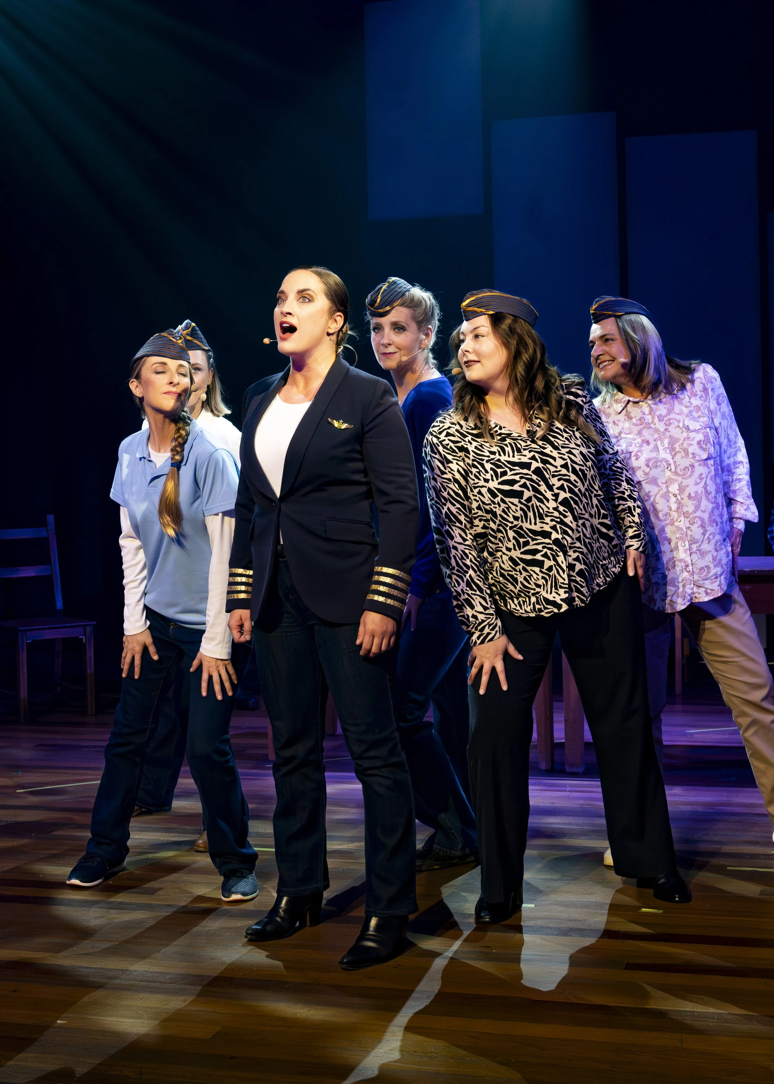A stage play featuring six women, with one woman in the center singing or acting passionately, dressed as a pilot with a black suit and gold stripes, surrounded by women in casual and business attire, some wearing pilot hats, under stage lighting.
