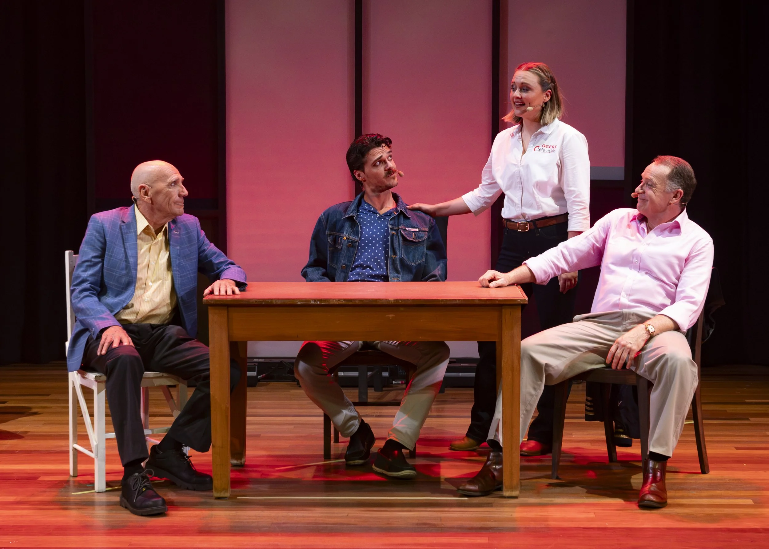 Stage scene with four actors: one woman standing and speaking, three men sitting around a wood table, all engaged in conversation, in a theater setting.